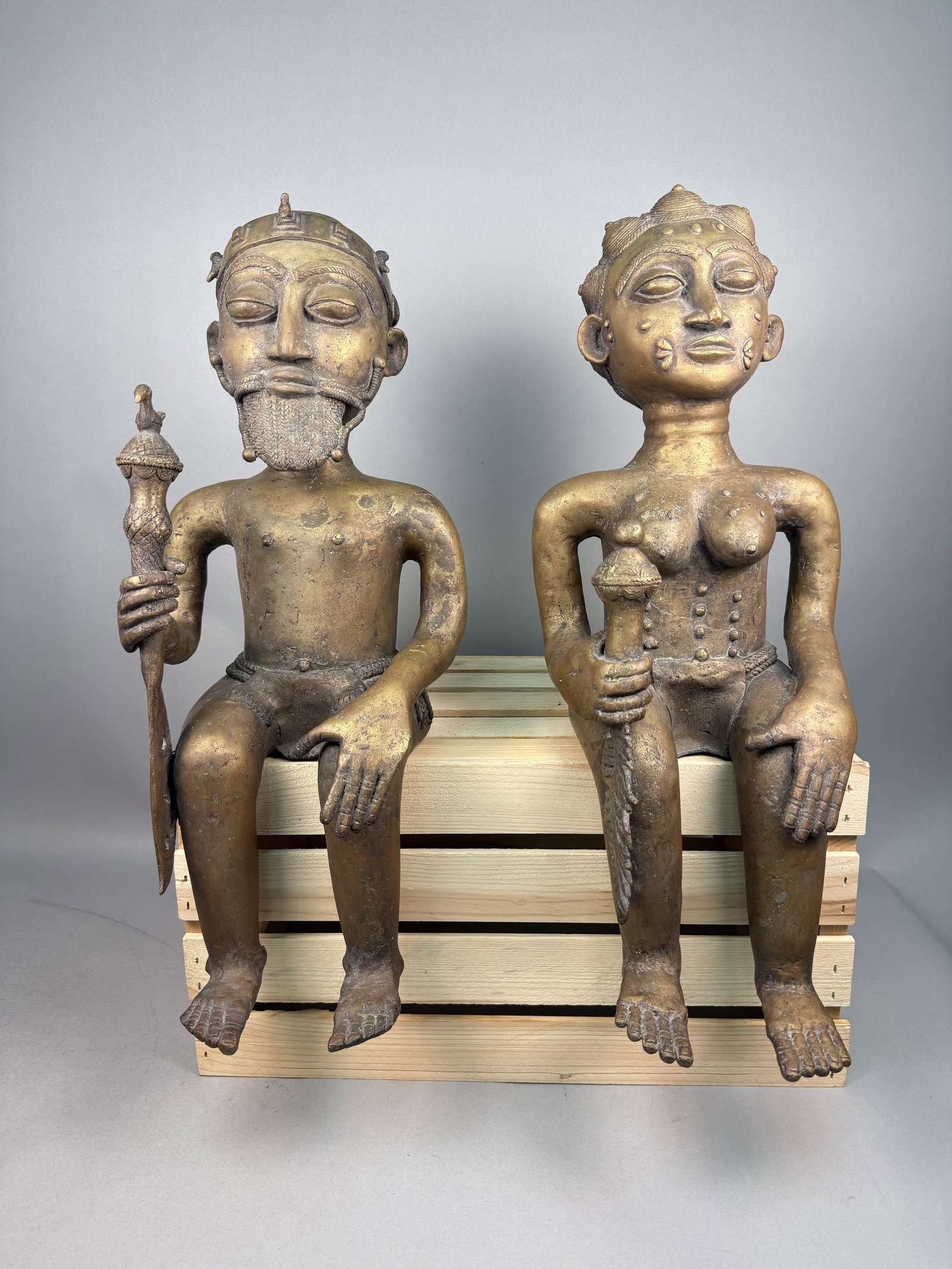 Large West African Cast Metal Seated Figures: A pair of large seated figurative sculptures, one male and one female, created in a West African style resembling casting traditions from Nigeria or Cameroon.The surfaces of both pieces show visible