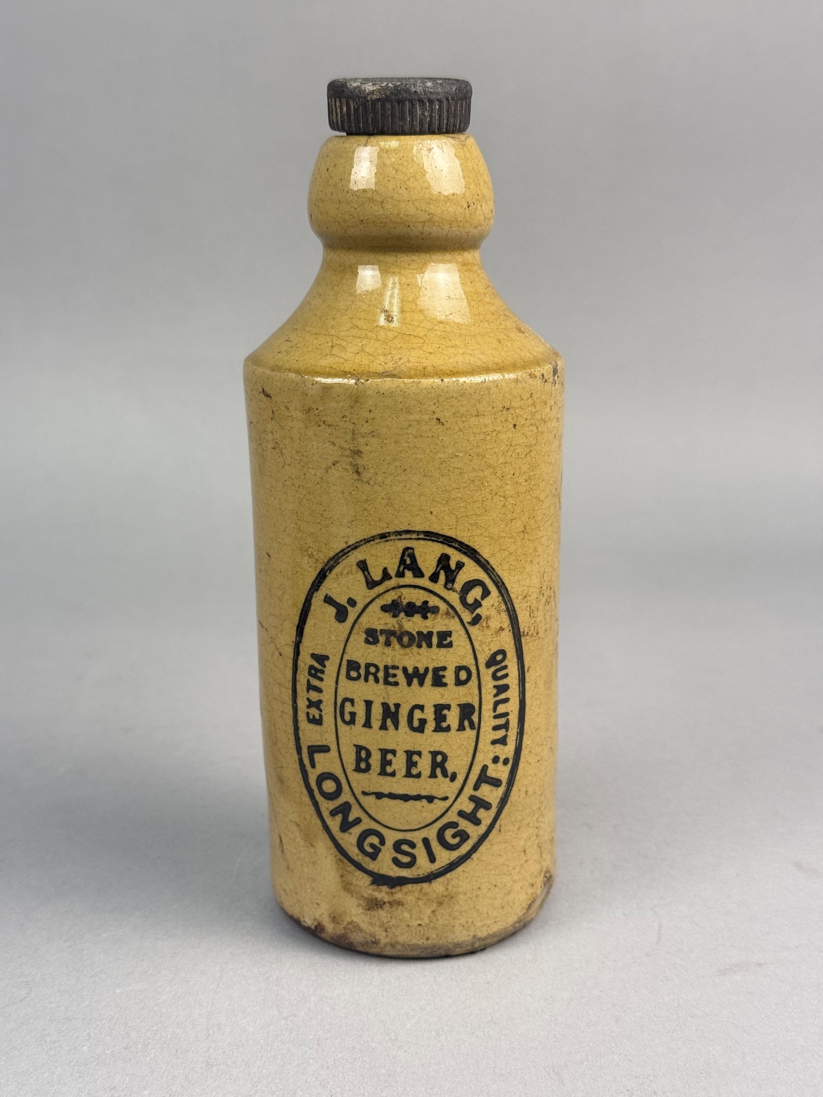 J. Lang Longsight Manchester Ginger Beer Bottle: This antique British stoneware ginger beer bottle is of a cylindrical bullet form with a bulbous neck containing an internal screw thread. The body is crafted from stoneware with a yellowish-tan glaze