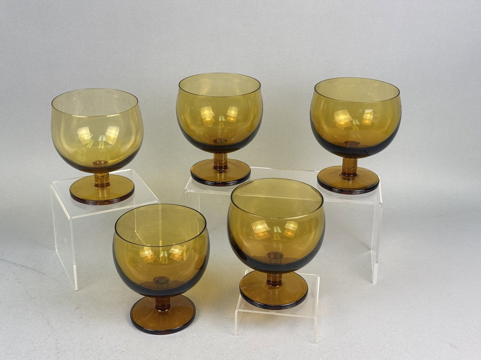 Five Mid-Century Amber Glass Short Goblets: A set of five mid-century modern short goblets constructed from translucent honey-amber colored glass. Each vessel features a wide, bulbous bowl with a subtly tapered rim, supported by a short cylindr