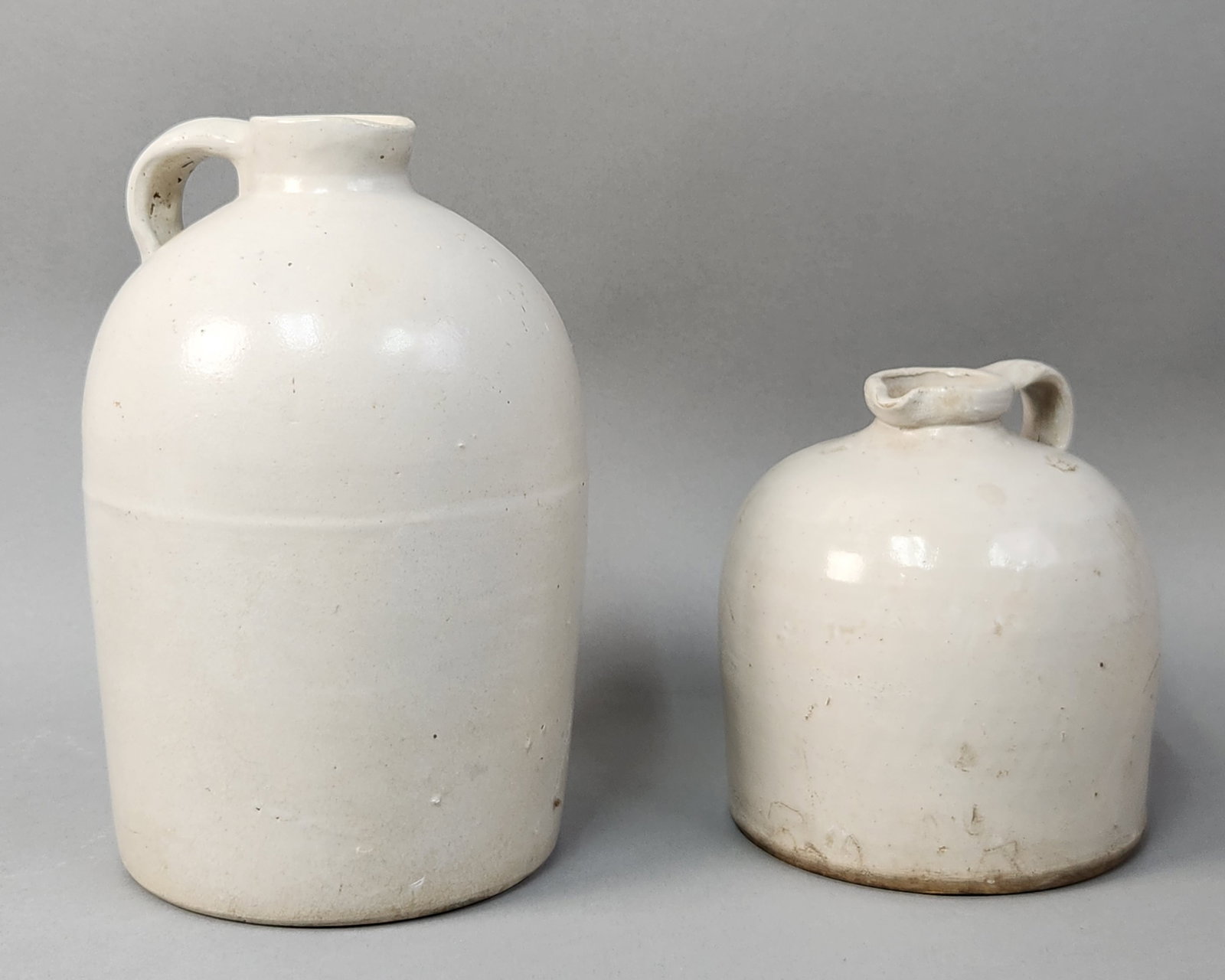 Bristol Glaze Stoneware Jugs: This pair of antique American stoneware. Both pieces are finished in a clean white Bristol glaze. The larger jug is a two-gallon capacity vessel and features an impressed circular maker mark on the ba