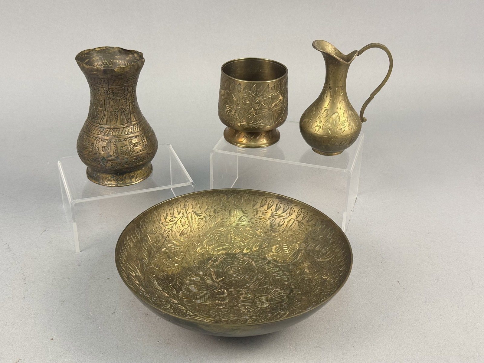 Four Indian Etched Brass Vessels: This group consists of multiple brass vessels featuring hand-etched and stippled decoration. The collection includes a large low bowl, a small vase, a footed cup, and a small pitcher with a handle. Th