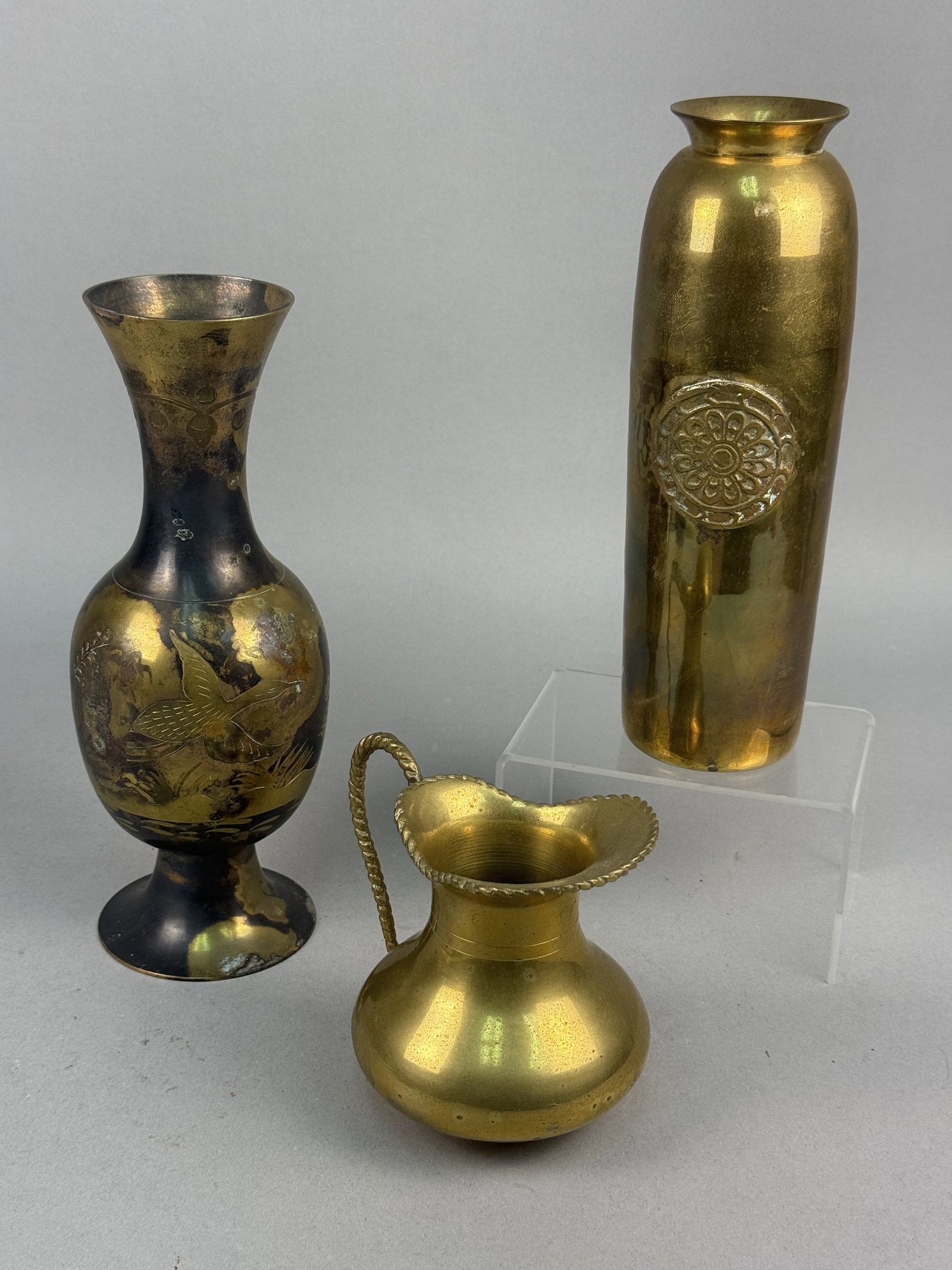A Trio of Decorative Brass Vessels: This lot consists of three decorative brass vessels. The first is a slender-necked vase featuring a flared rim and a pedestal base. The exterior has a blackened, patinated finish with etched decoratio