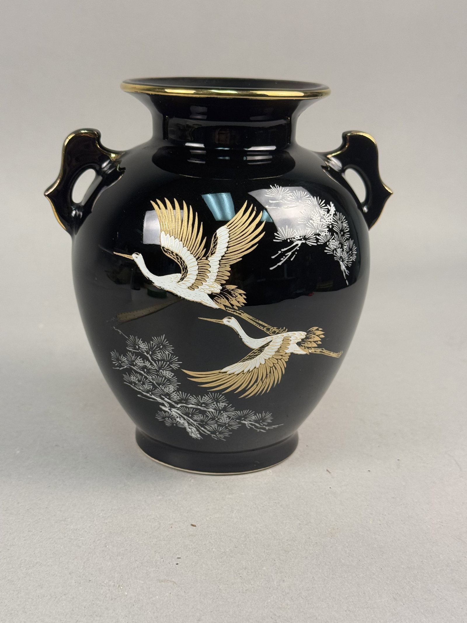 Japanese Black Glazed Porcelain Vase Crane Motif: A Japanese porcelain export vase featuring a bulbous body, pedestal base, flared rim, and two scrolled handles. The vessel is finished in a high-gloss black glaze with gilt trim along the rim and hand