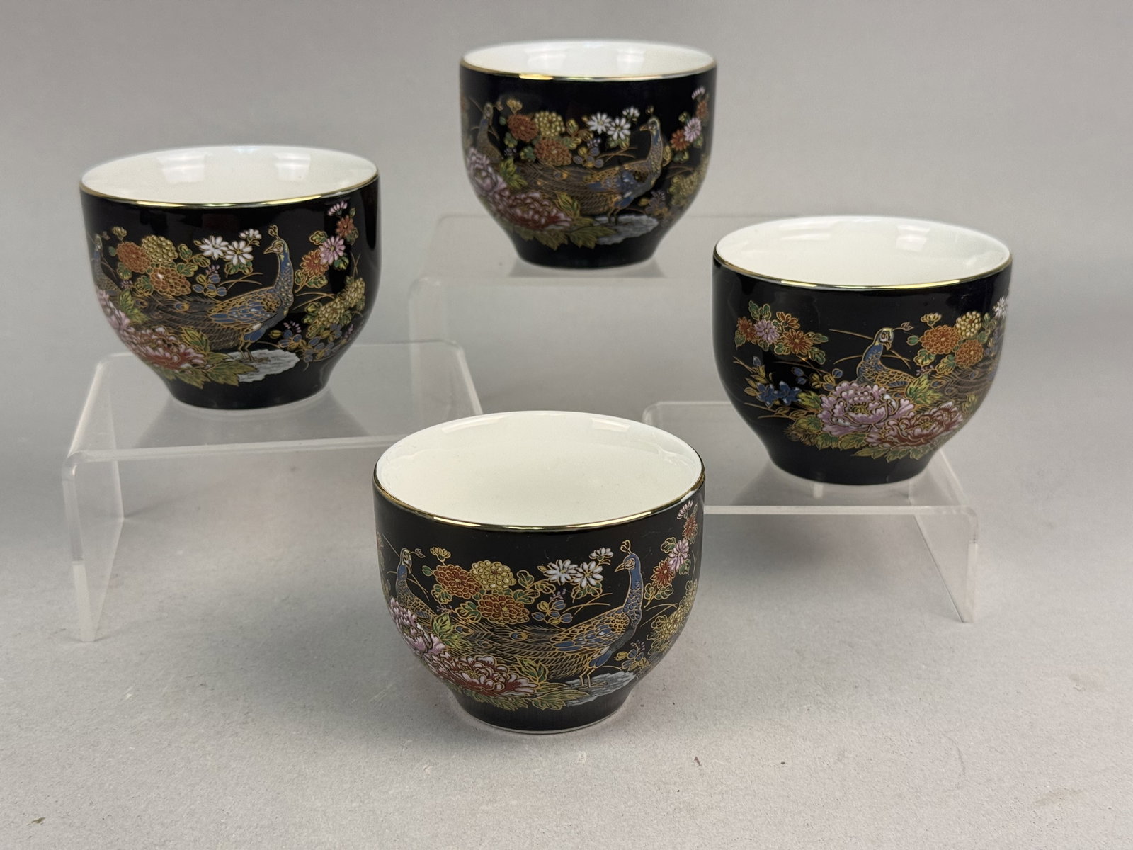 4 Japanese Porcelain Peacock Sake or Tea Cups: This set of four porcelain vessels is suitable for use as tea or sake cups. Each item features a polished black glazed exterior ground embellished with overglaze polychrome enamels and heavy gold outl