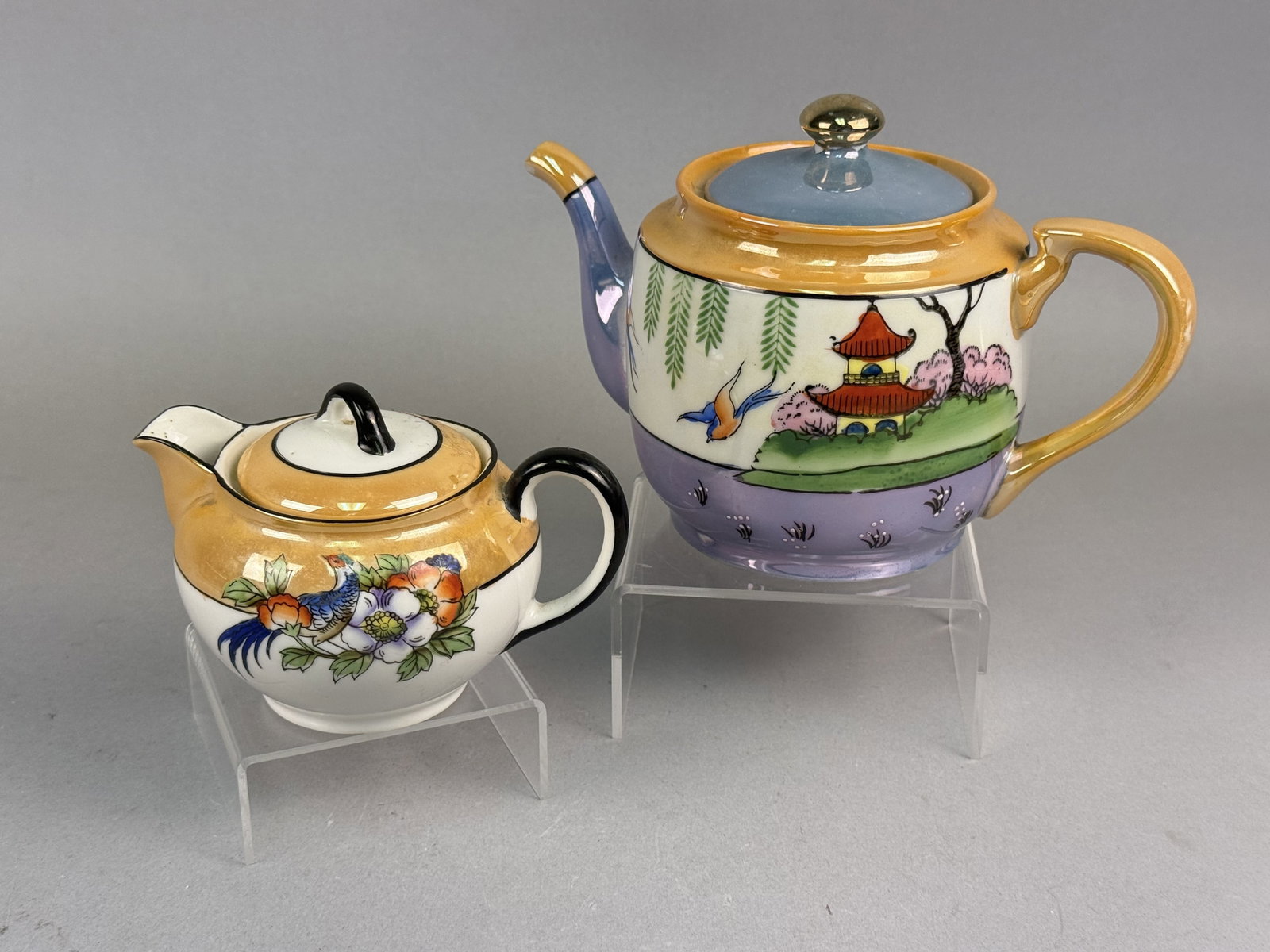Japanese Lusterware Noritake Creamer & Teapot: A two-piece set of Japanese hand-painted porcelain featuring iridescent luster glazes in orange, lavender, and blue tones. The teapot is designed with a rounded body and decorated with a pastoral land