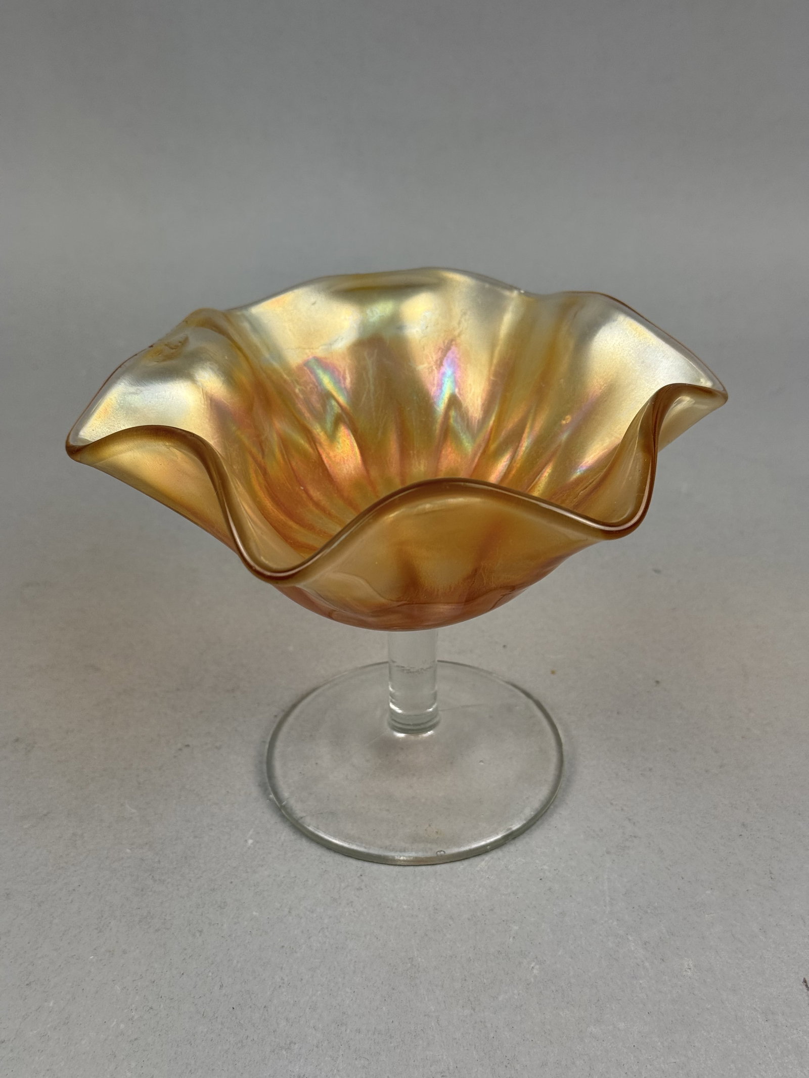 Marigold Carnival Glass Ruffled Compote: This lot consists of a mold-pressed carnival glass compote featuring a bowl with a marigold iridescence. The finish displays gold and peach tones with subtle purple and pink highlights. The bowl is