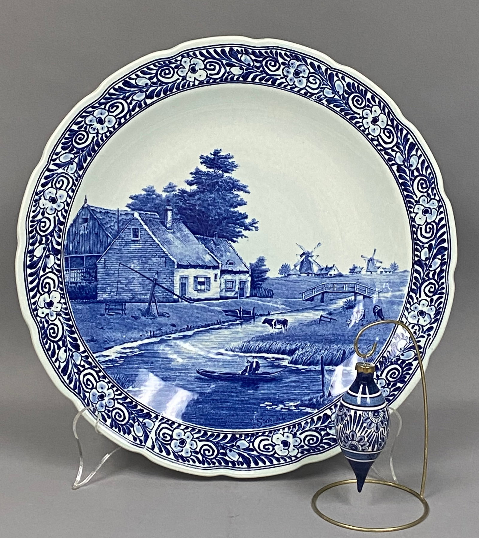 2 Pieces of Delft Pottery Inc a Royal Sphinx Maastricht Charger: This lot consists of a large blue and white ceramic wall charger and a coordinating hand-painted ornament. The earthenware charger by Royal Sphinx Maastricht features a scalloped edge and a central pa