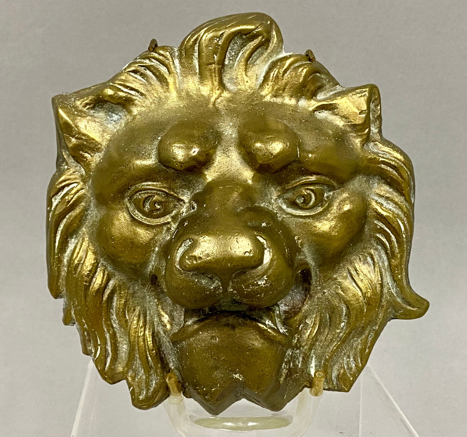 Early Cast Brass Lion Head Decorative Mount: A hollow-cast brass decorative mount or plaque depicting a lion's head in relief. The piece features stylized Neoclassical details, including a prominent wavy mane, heavy brow, and deep-set eyes. The