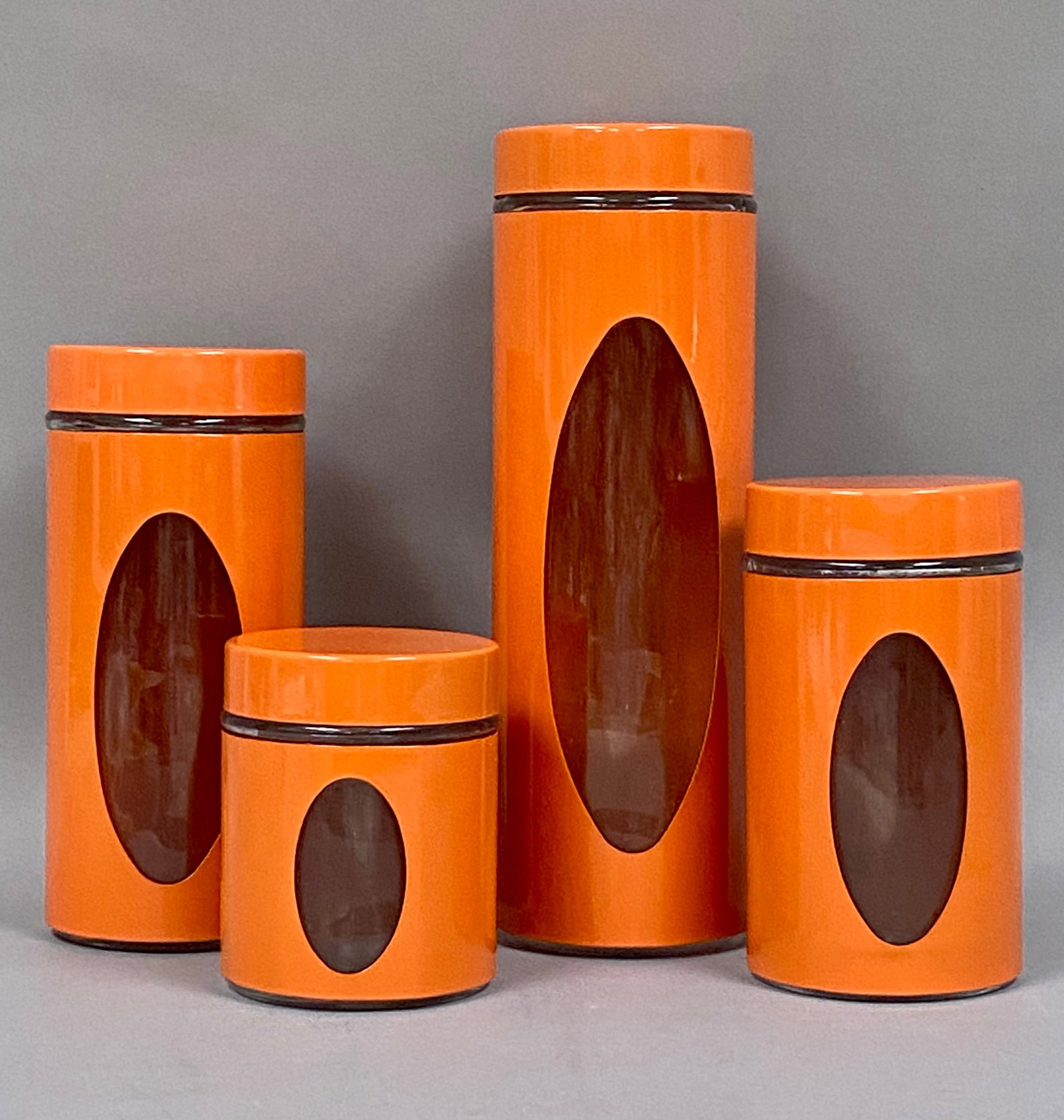 MCM Style Glass & Orange Metal Kitchen Canisters: A set of four kitchen canisters of graduated heights, each constructed from a clear glass jar encased within a vibrant orange-finished metal sleeve. The sleeves feature a large, vertical oval viewing 