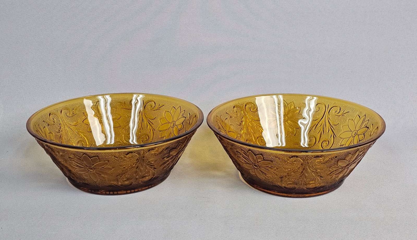Pair of Tiara Sandwich Amber Bowls: A pair of translucent amber pressed glass bowls in the Sandwich pattern by the Indiana Glass Company. The vessels are decorated with pressed floral medallions, scrolling vines, and a stippled ground. 