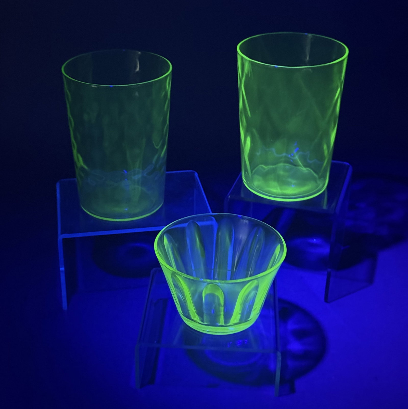 Trio of Green Uranium Glass: This trio of green glass includes two tumblers with a honeycomb optic pattern and a flared bowl featuring vertical fluting. All pieces demonstrate characteristic bright green fluorescence under