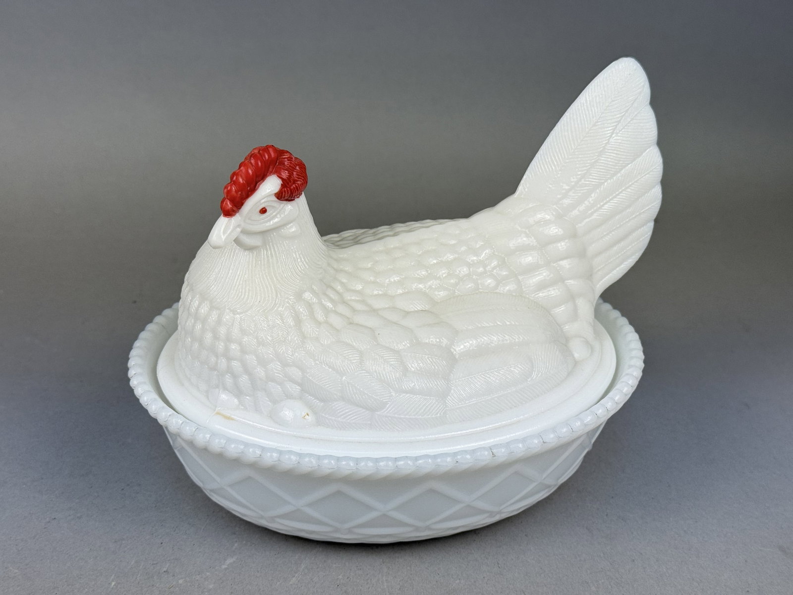 Tiara Exclusives Milk Glass Hen on Nest: This Tiara Exclusives covered dish is fashioned in the classic hen on nest form, crafted from opaque white milk glass. The lid features a nesting hen with detailed molded feathers and red