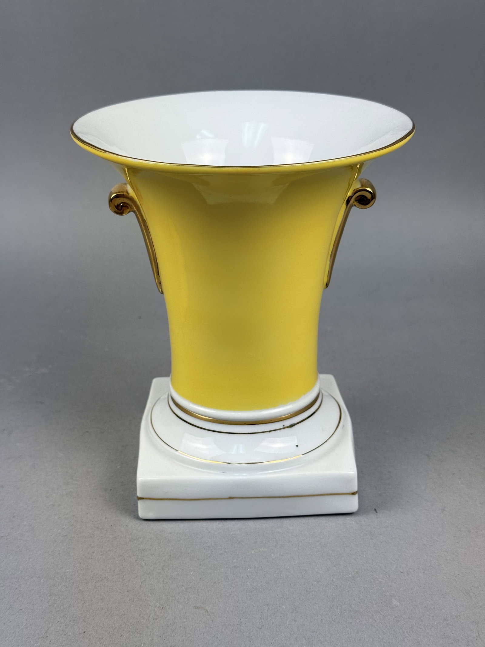 Yellow Ground Gilded Porcelain Urn: This porcelain urn-form vase features a vibrant canary-yellow glazed body accented with gilded scroll handles. The flared rim and tiered circular base are finished with hand-applied gold bands atop a