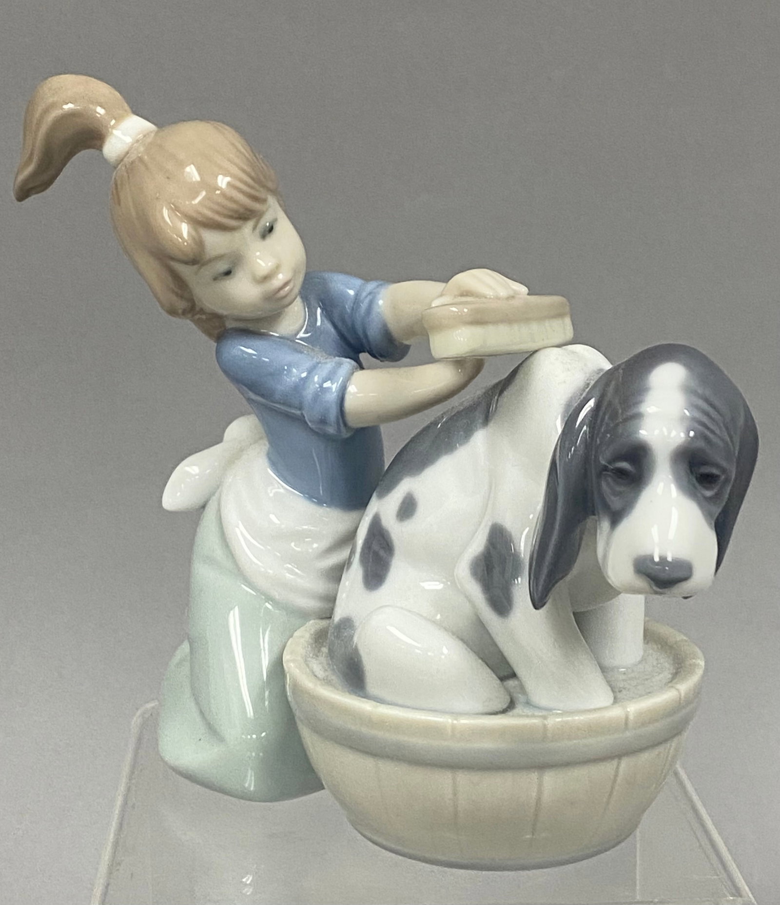 Lladro Bashful Bather, Number 5455: This Lladro glazed porcelain figurine is titled Bashful Bather, model number 5455. The piece depicts a young girl with a light brown ponytail kneeling as she gives a bath to a spotted, floppy-eared do