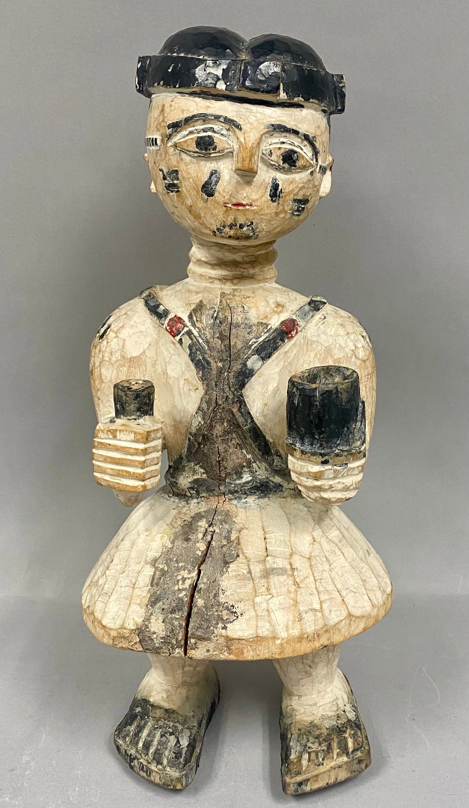 West African Carved Wood Standing Figure: The figure is depicted wearing a black cap and a wide, tiered skirt. A cross-strap harness with red circular accents is carved across the chest and back. The figure holds two cylindrical vessels, one