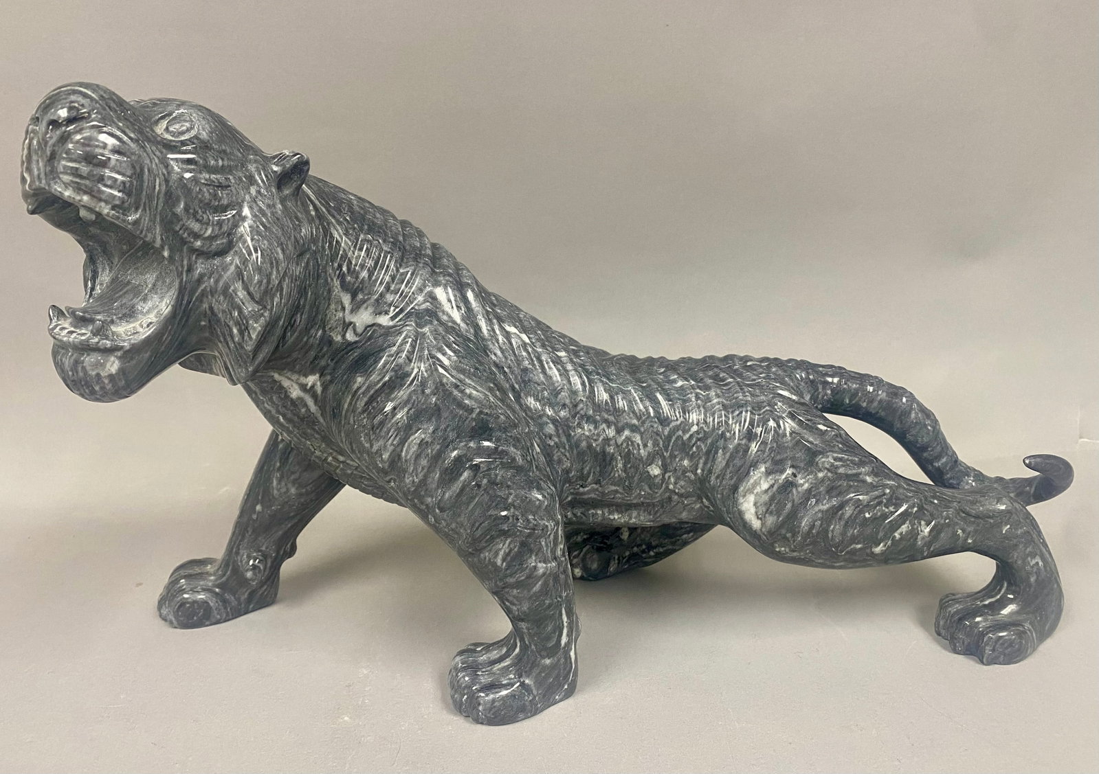 Large Carved Marbled Stone Roaring Tiger Sculpture: A large decorative sculpture depicting a tiger in a dynamic prowling pose with its head raised and mouth open in a roar. The piece is carved from a grey and white marbled stone with a polished finish 