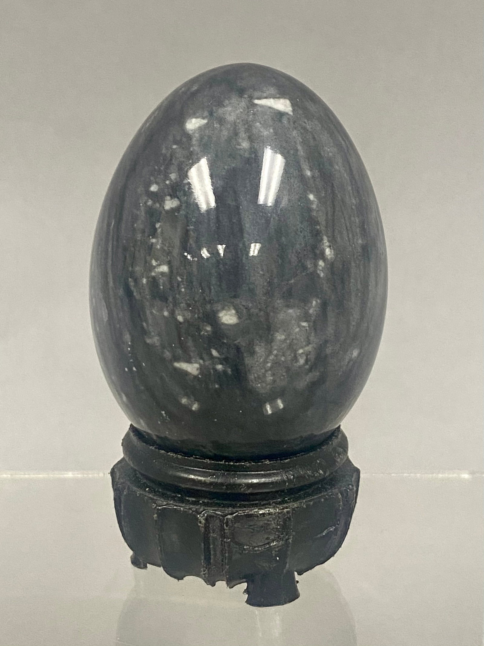 Polished Mottled Gray Stone Egg on a Carved Wooden Stand: A polished decorative egg crafted from a mottled dark gray and blue-gray natural stone featuring white inclusions throughout the mineral. The stone exhibits a high-gloss finish. The egg is supported b