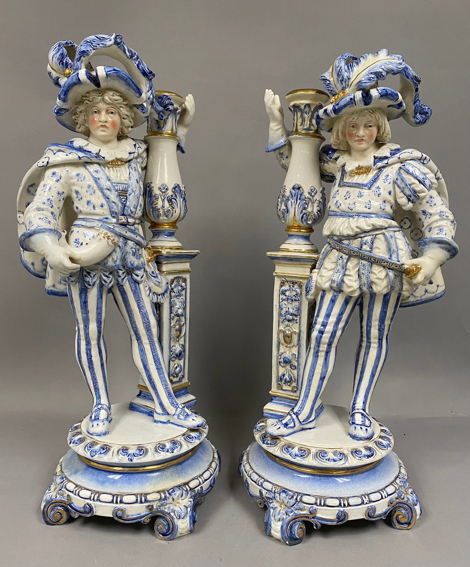 Ernst Bohne Sohne Porcelain Figural Candelabra: A pair of Ernst Bohne Sohne glazed porcelain figural candelabra bases modeled as Renaissance pages in blue and white. Each figure leans against an architectural pedestal finished with gilded accents