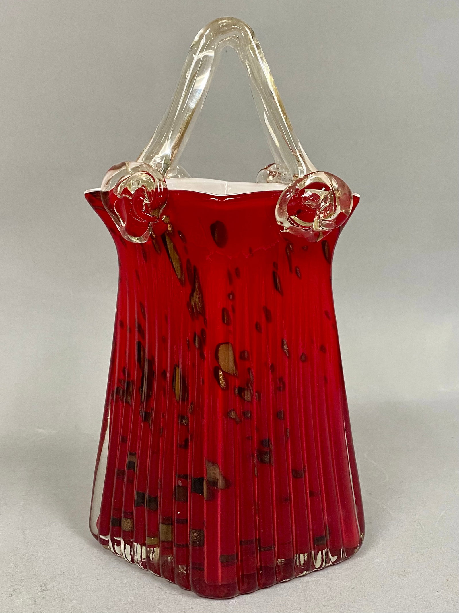Ruby Red Aventurine Glass Purse: This hand-blown cased glass vase is crafted in the form of a handbag with a vibrant ruby red exterior and an opaque white interior. The body features vertical optic ribbing and gold aventurine flecks