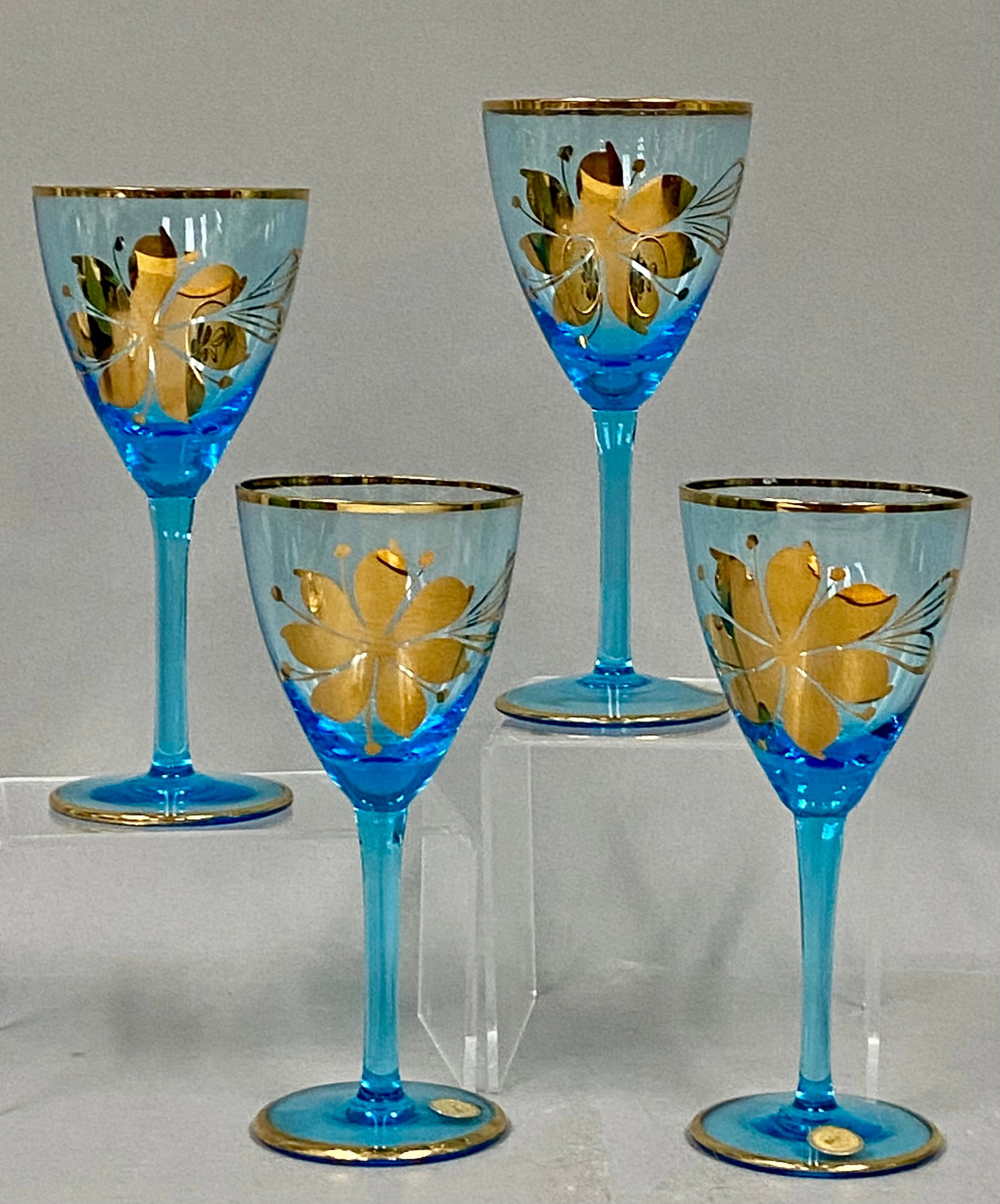 Set of Romanian Blue Glass Goblets: A set of four Romanian aqua blue glass goblets decorated in a stylized gold floral pattern. Each vessel features a vibrant turquoise hue accented by metallic gilding at the rims and circular bases.