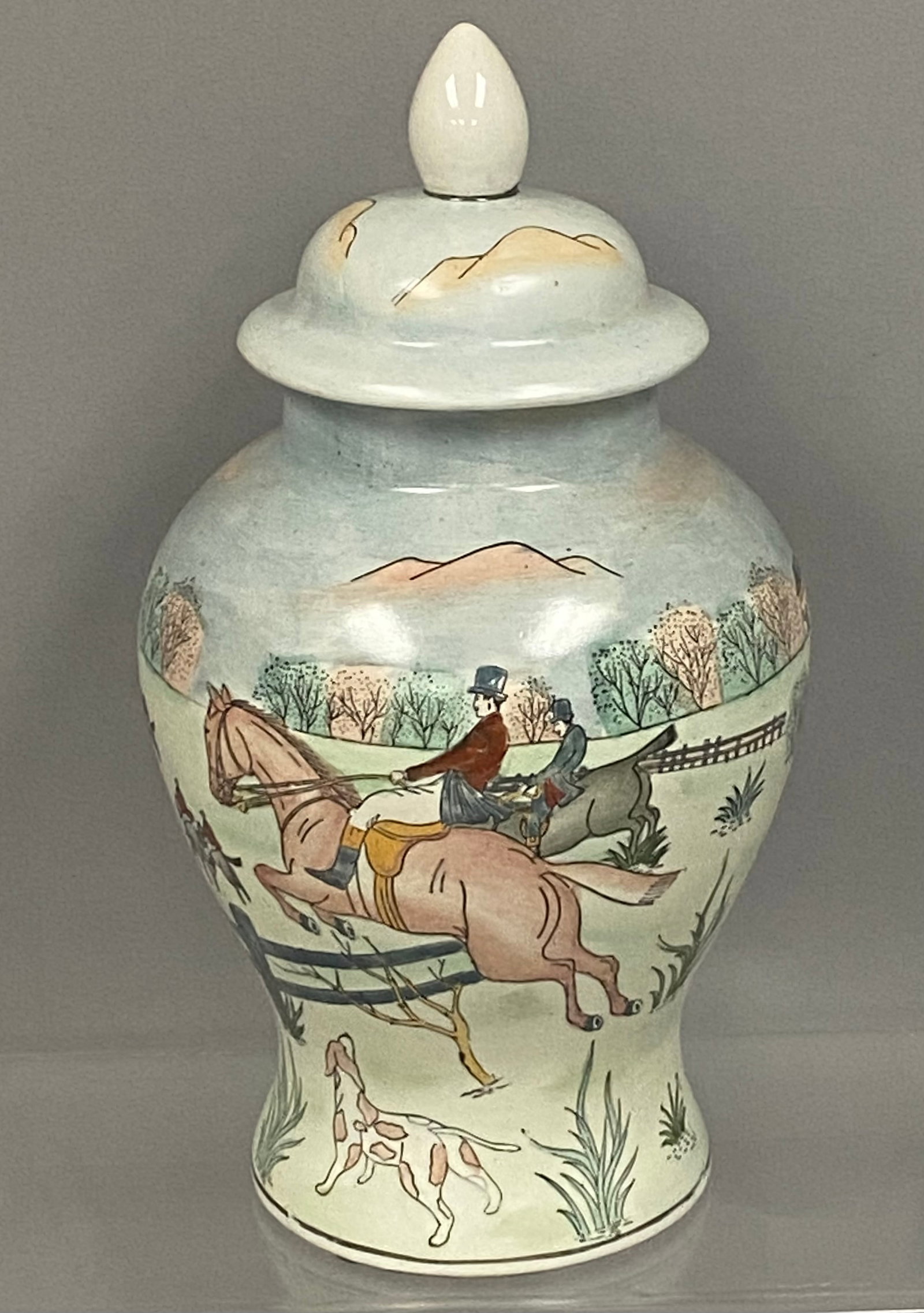 Lidded Ceramic Fox Hunt Ginger Jar: This lidded ceramic ginger jar features an equestrian fox hunting scene with riders jumping a post-and-rail fence. The landscape is rendered in soft color washes and includes a spotted hound, rolling