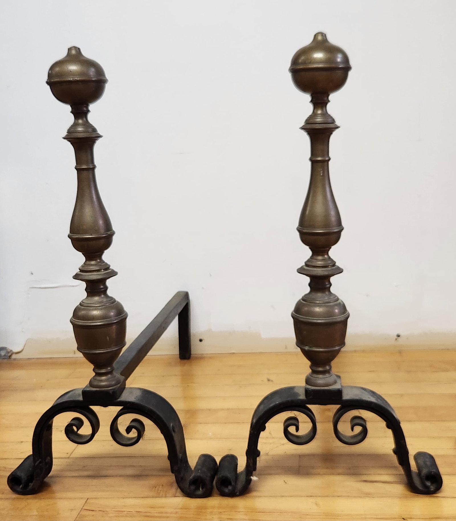Pair Turned Brass and Iron Andirons: This pair of andirons features multistage turned brass uprights terminating in ball finials. Height: 21.5 inches. The pair is in good structural condition. The brass displays a natural aged patina