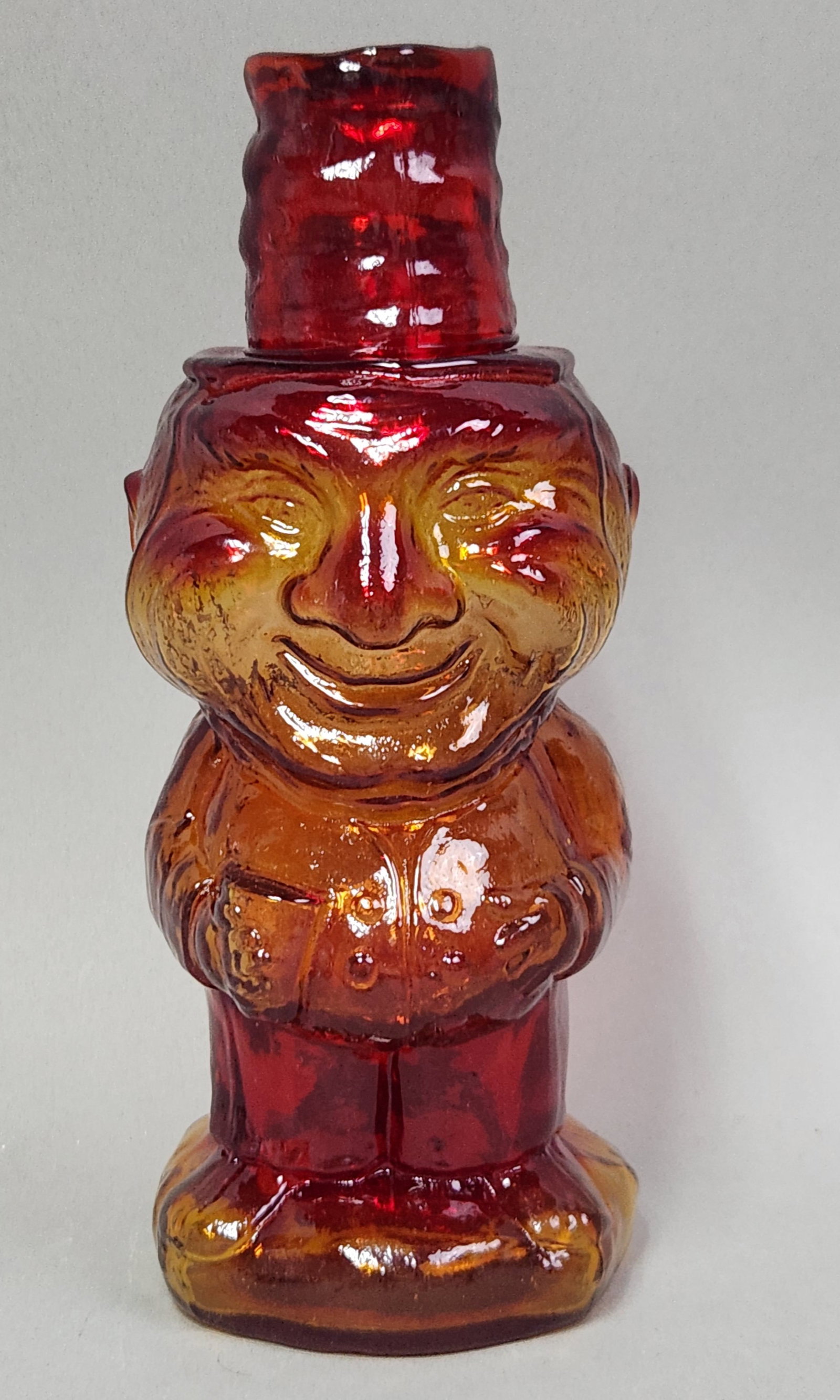 Indiana Glass Jolly Mountaineer Decanter: Amberina Indiana Glass Jolly Mountaineer figural decanter complete with a coordinating top hat stopper. Height: 10.5 inches. A short hairline crack is present at the interior rim. The surface shows