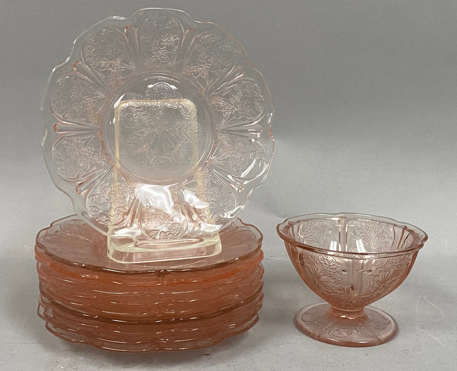 Jeannette Pink Cherry Blossom Glass Collection: A collection of seven pieces of pink translucent Depression-era glass in the Cherry Blossom pattern manufactured by the Jeannette Glass Company. The set includes ten plates featuring a central molded
