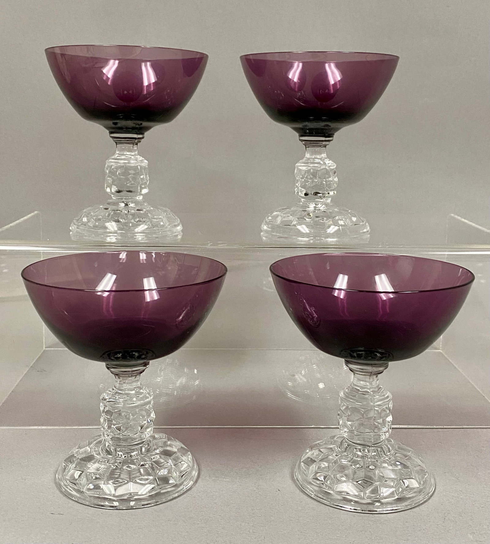 Four Amethyst Stemware Glasses: A set of four glass coupes featuring deep amethyst purple bowls. Each piece is designed with a heavy, molded diamond-point pattern on the clear stem and tiered circular base. The construction shows