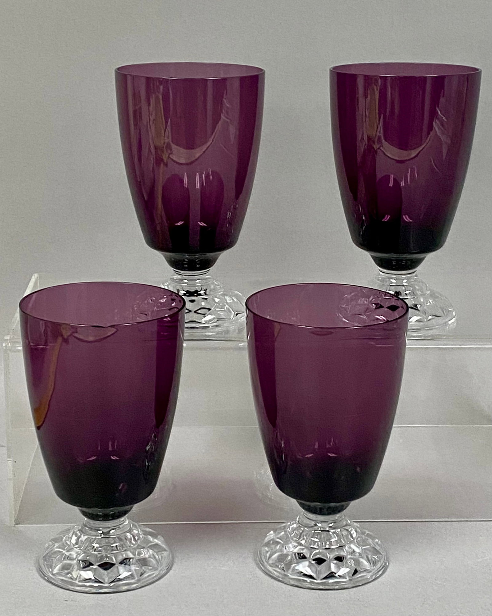 Four Amethyst Goblets: This set of four goblets features deep amethyst purple bowls with plain rims. Each vessel sits on a short, clear faceted stem and a circular foot decorated with a diamond-point cut pattern. The