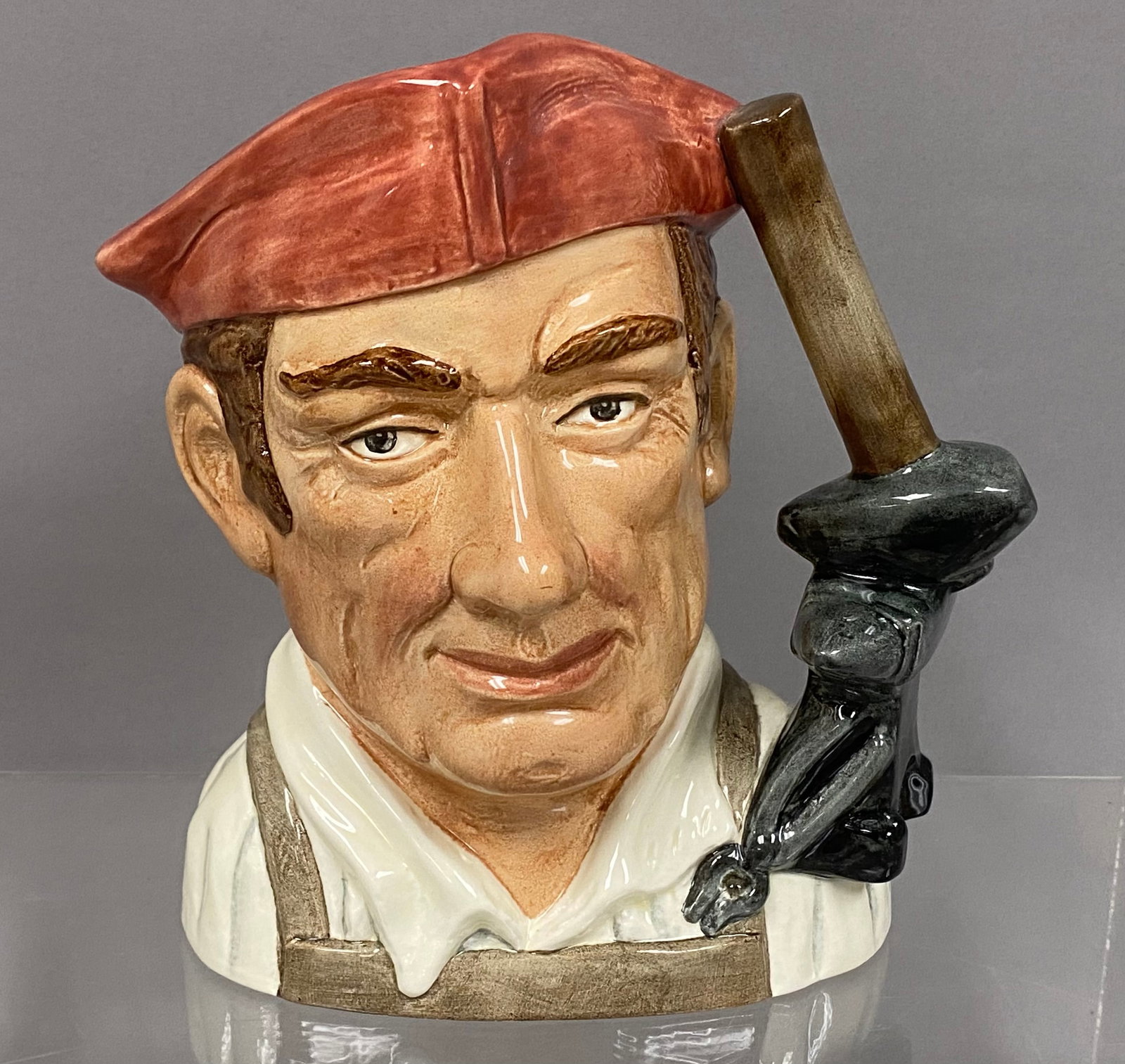 Royal Doulton Blacksmith Character Jug: This Royal Doulton glazed ceramic character jug depicts a blacksmith from the Character Jugs from Williamsburg series. The figure is modeled with a red peaked cap and a ponytail secured with a black