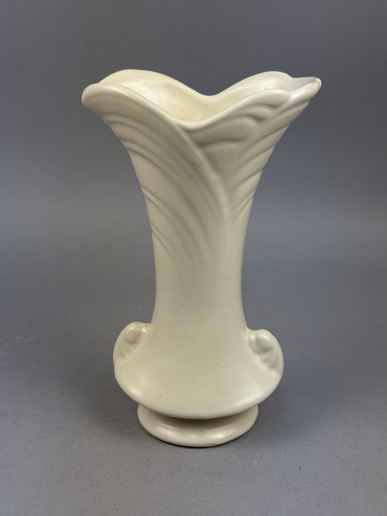 Shawnee Pottery Cream Leaf Vase: This Shawnee Pottery vase is crafted from glazed earthenware with a monochrome cream-colored satin finish. The vessel features a flared ruffled rim with molded leaf-like vertical ribbing and two