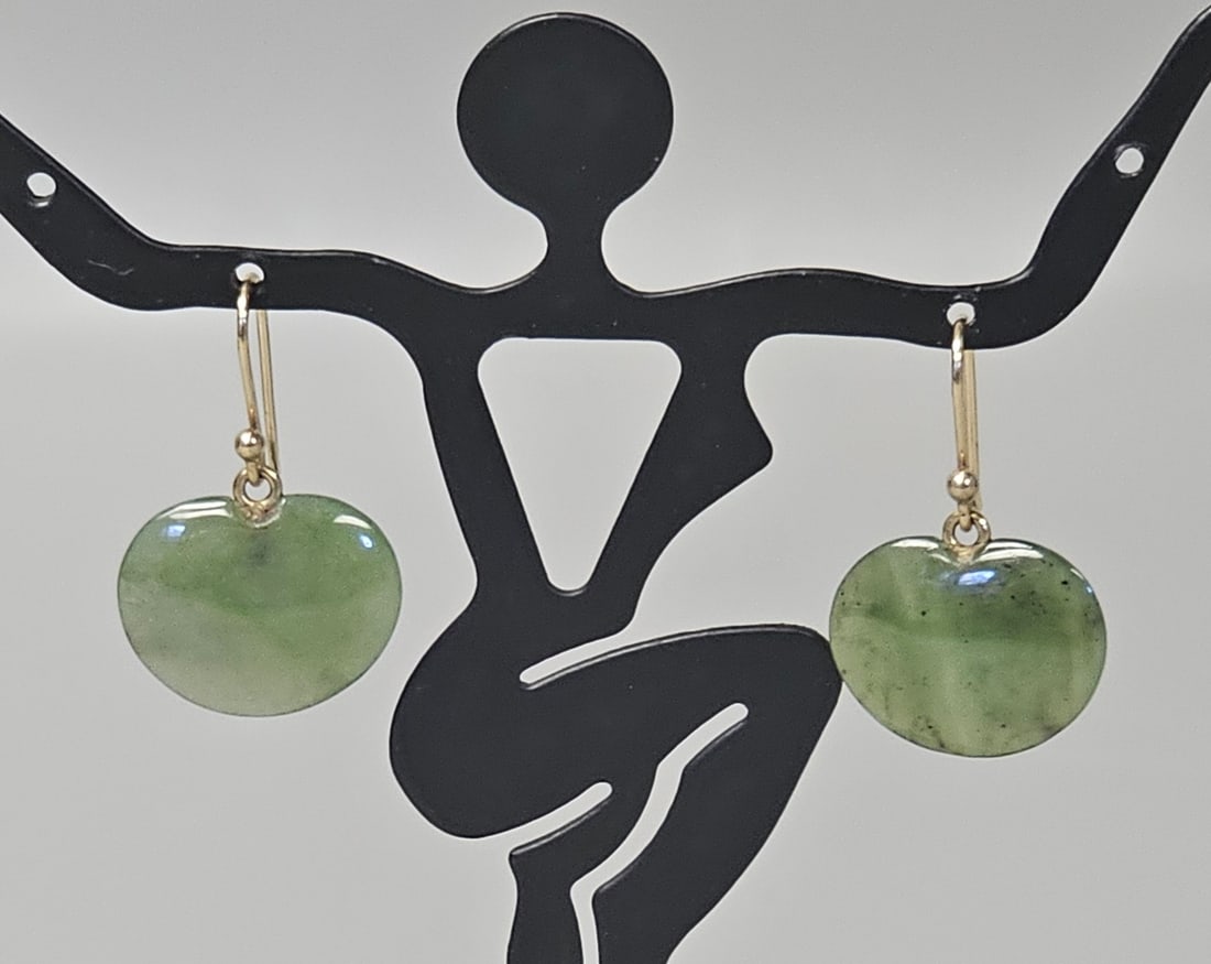 14K Gold Nephrite Jade Earrings: This pair of drop earrings features polished, heart-shaped nephrite jade cabochons displaying characteristic green mottling and dark "spinel" inclusions. Each jade drop is suspended from yellow gold s