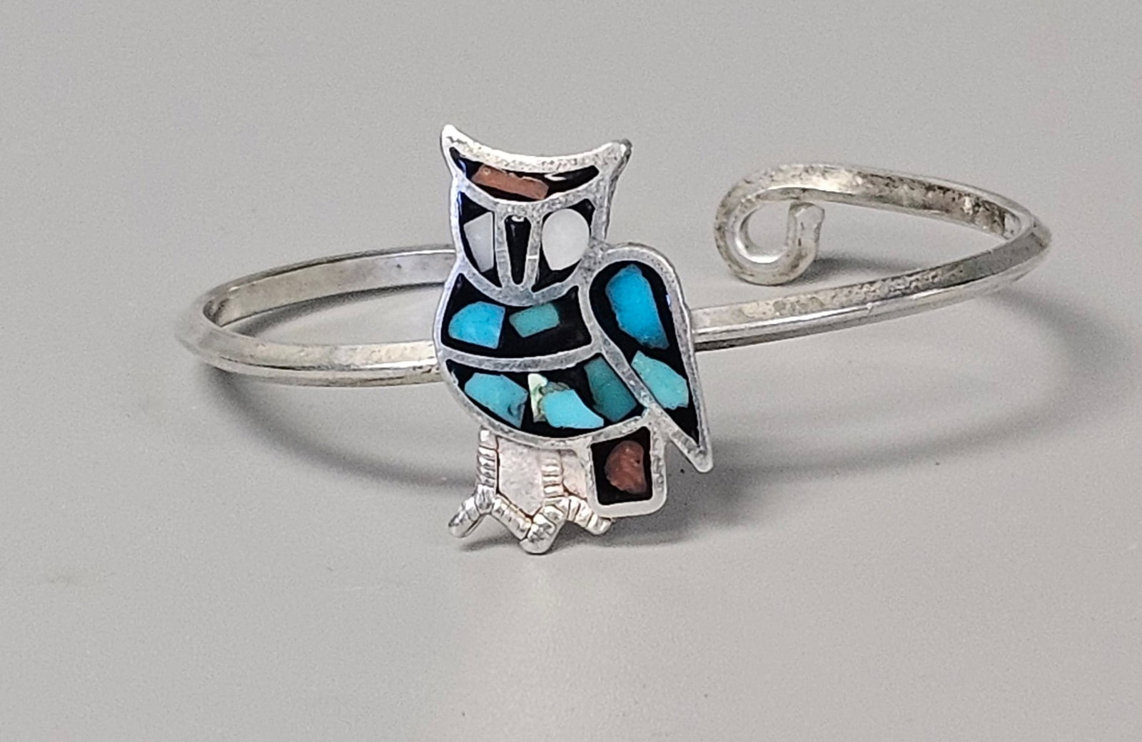 Zuni-Style Inlay Owl Silver Bracelet 10.0 Grams: This handcrafted cuff bracelet features a traditional Zuni-style owl motif rendered in stone-to-stone channel inlay. The plaque is set with turquoise, mother-of-pearl, jet, and coral, mounted on a sil
