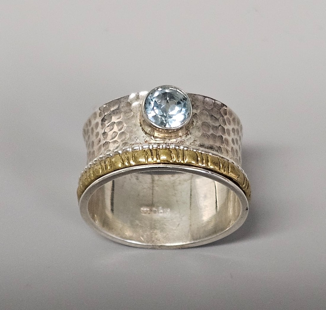 Sterling Silver and Gemstone Spinner Ring 6.2 Grams: This size 11 mixed-metal spinner ring features a wide, hammered .925 sterling silver band accented by a rotating gold-toned textured element. A round, bezel-set faceted light blue gemstone serves as