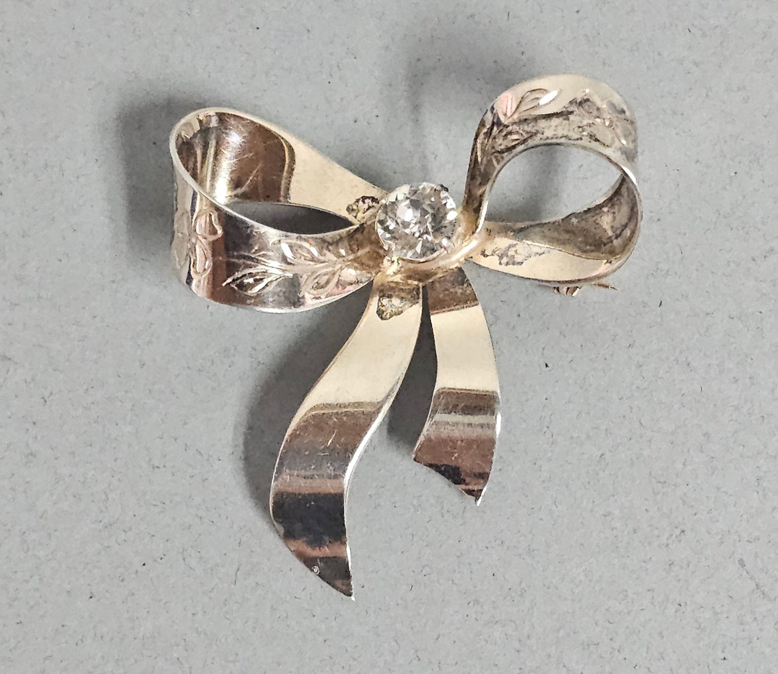 Sterling Vermeil Retro Bow Brooch 8.1 Grams: This mid-20th-century Retro-period brooch is fashioned as a three-dimensional bow with engraved foliate motifs and a central colorless faceted stone. It is constructed of sterling silver with a gold
