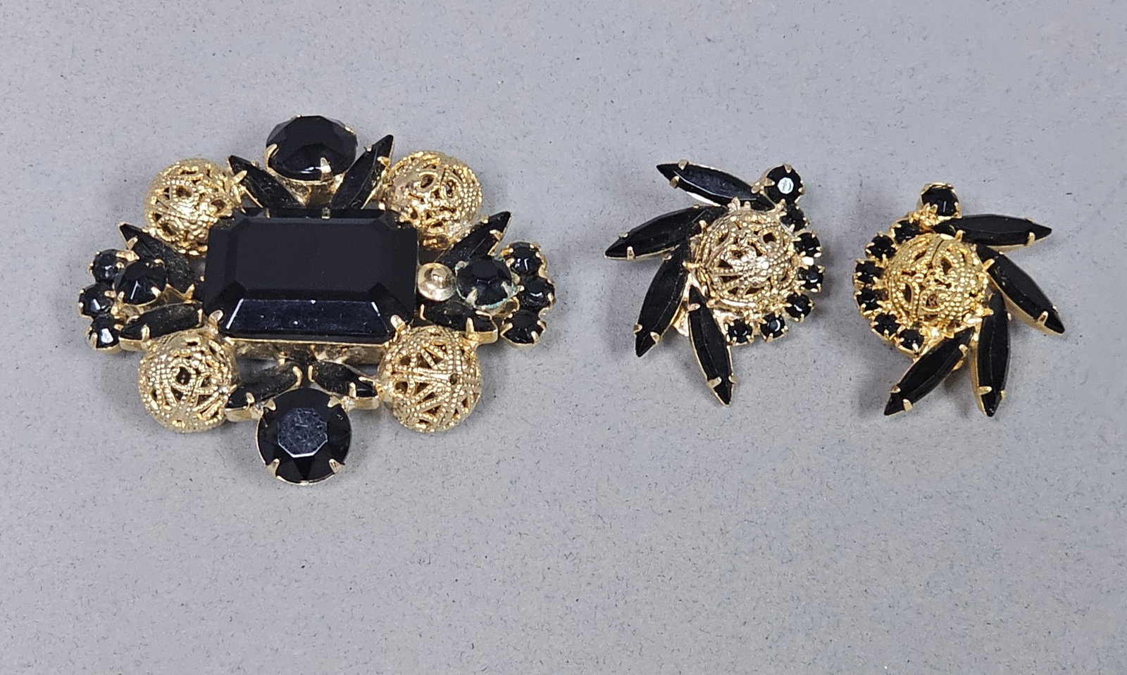 Juliana Black Glass Demi-Parure: This Juliana demi-parure by DeLizza and Elster consists of a brooch and matching clip-on earrings crafted in gold-tone metal. The set features large emerald-cut black glass stones accented by navette-