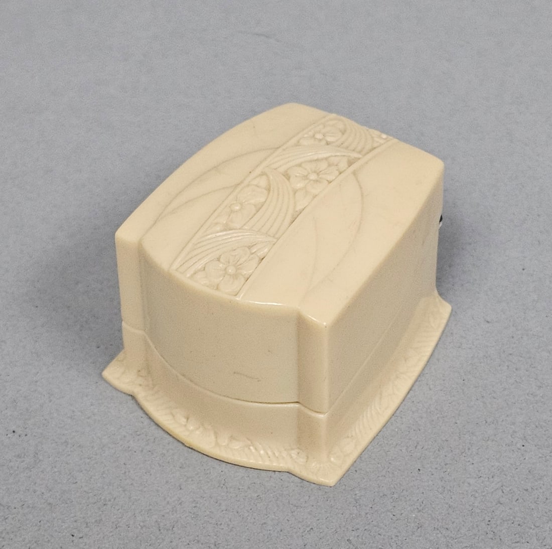Art Deco Floral Celluloid Ring Box: This Art Deco jewelry presentation box is constructed from cream-toned molded celluloid featuring stylized floral bands and sweeping geometric lines. The stepped form rests on a flared base with a
