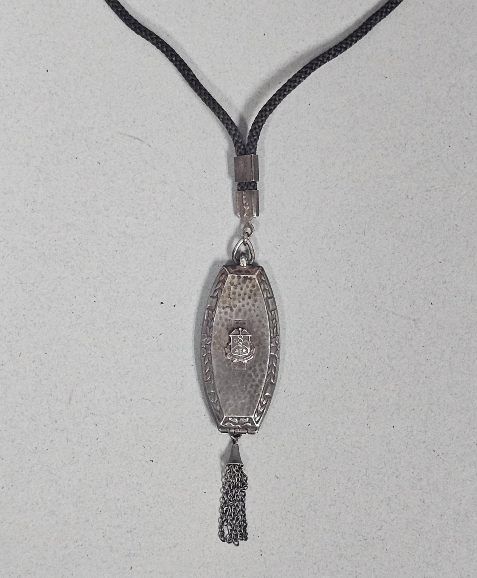 Art Deco Sterling Medical Locket 8.1 Grams: This navette-shaped Art Deco locket is crafted from hand-hammered sterling silver and features an applied shield-shaped caduceus medical crest and a chain tassel. The 8.1-gram pendant is suspended