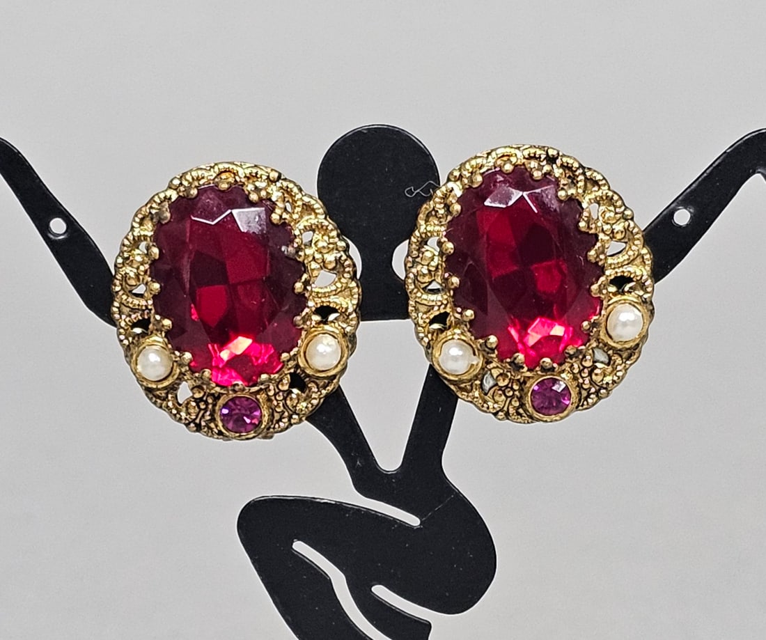 West German Red Glass Earrings: A matched pair of West German vintage costume clip-on earrings featuring ornate gold-tone filigree settings. Each earring centers an oval-cut faceted red glass stone, accented by a single round pink