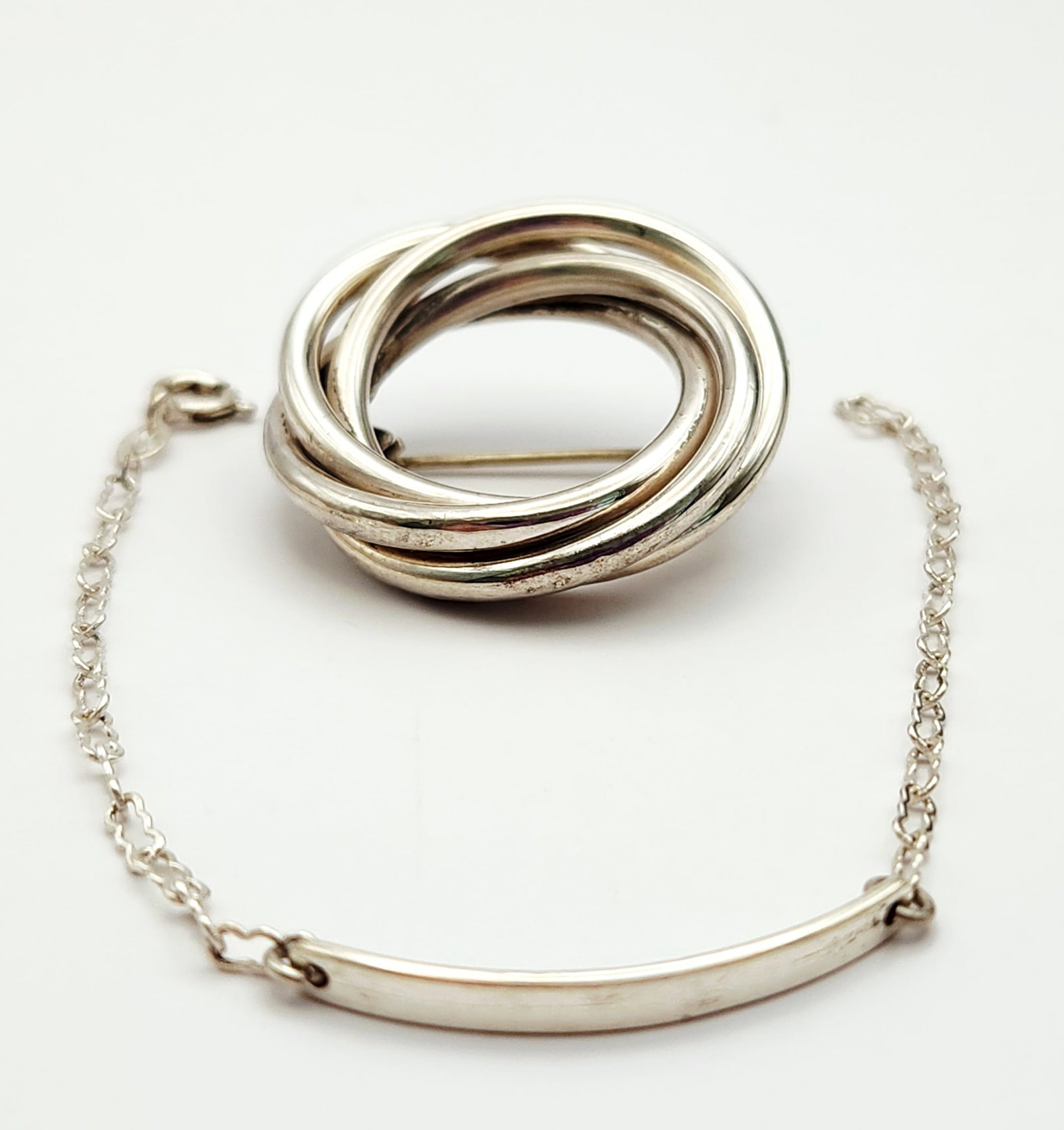 Sterling Pin & Bracelet 11.2 Grams: This lot features a James sterling silver brooch with intertwined rings and a 925 silver chain bracelet with a curved identification bar. The total weight for both pieces is 11.2 grams. Bracelet is 6"