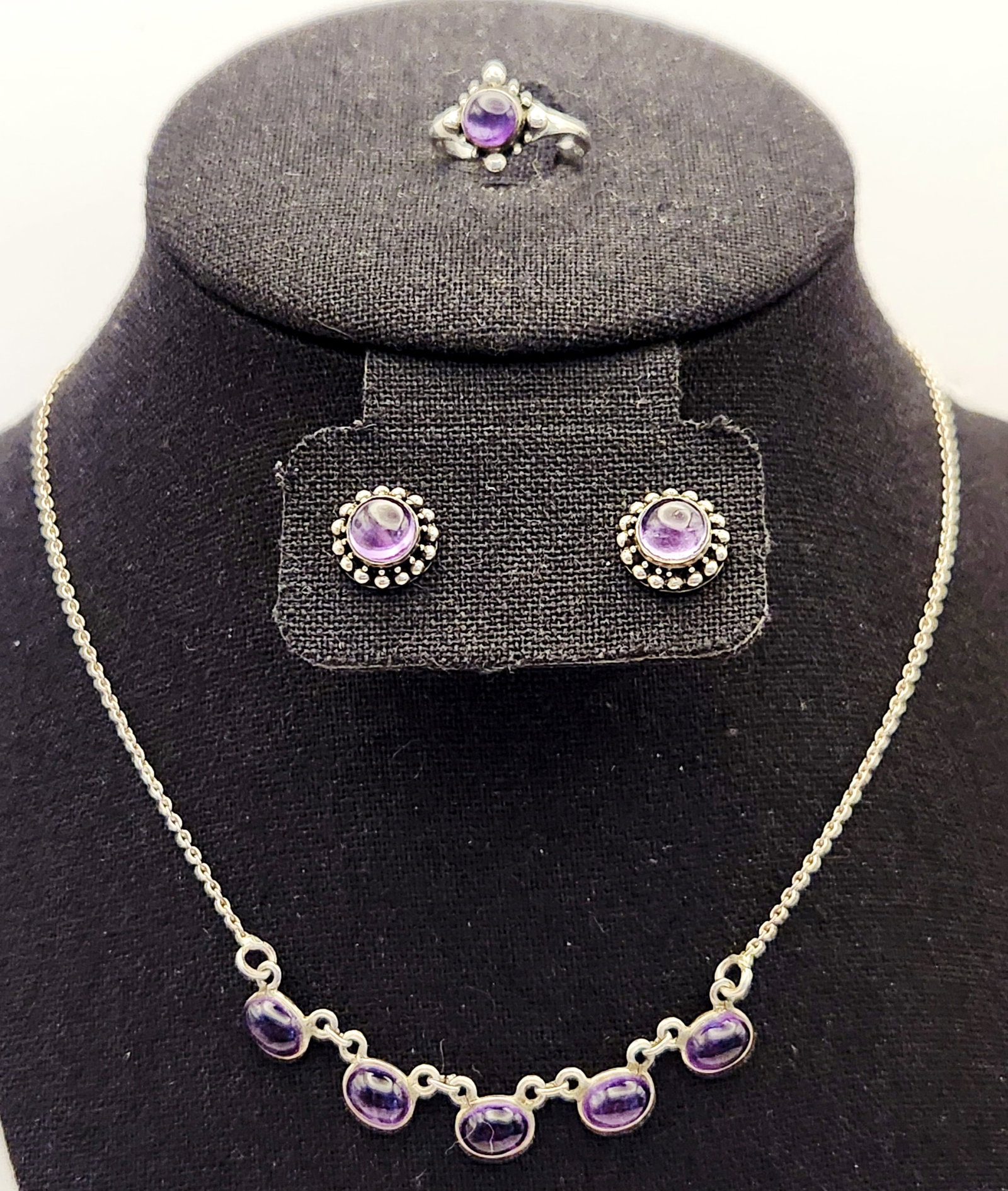 Sterling Amethyst Jewelry 10.2 Grams: A group of jewelry comprising a necklace, a pair of stud earrings, and a ring crafted in sterling silver and set with bezel-set purple amethyst cabochons. Total weight: 10.2 grams Ring size: 6.25
