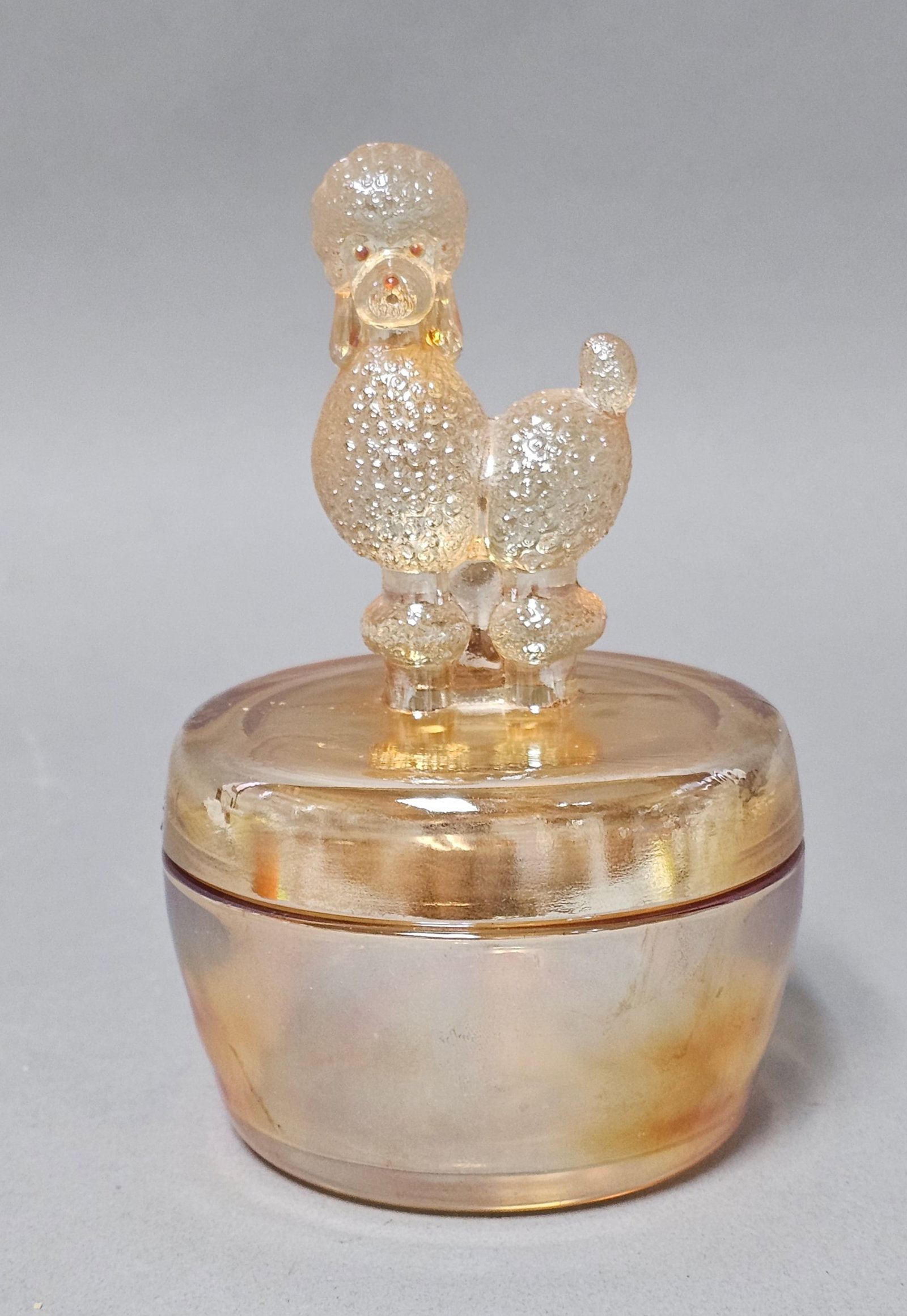 Jeannette Marigold Carnival Glass Poodle Jar: This two-piece powder jar is crafted in marigold carnival glass by the Jeannette Glass Company. The lid features a molded finial in the form of a poodle with a textured stucco clip, while the base is 