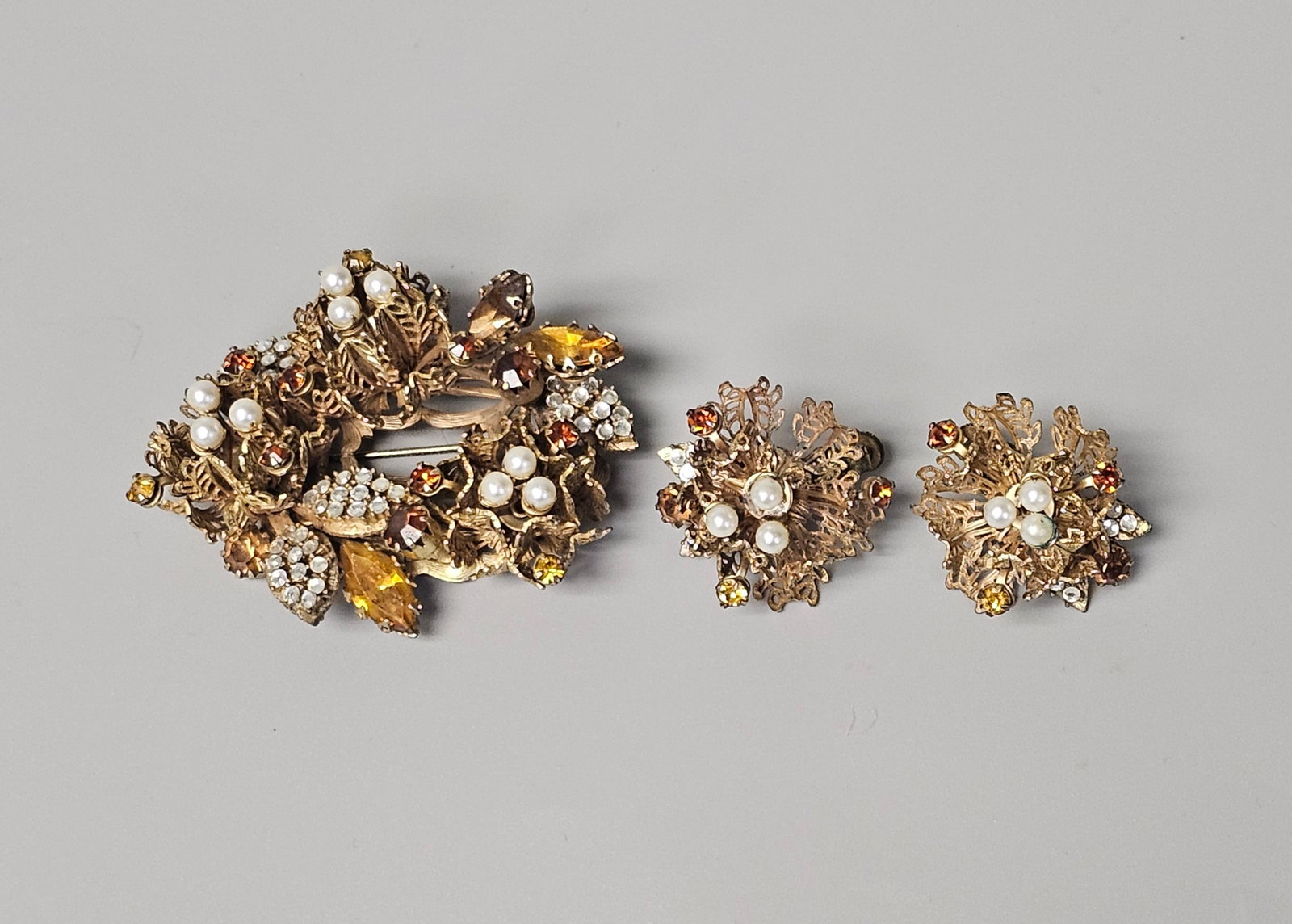 Vendome Mid-Century Gilt Costume Demi-Parure: A Vendome mid-century demi-parure featuring an ornate brooch and matching clip-on earrings. The pieces are constructed from gold-tone filigree metal set with amber and topaz-colored glass