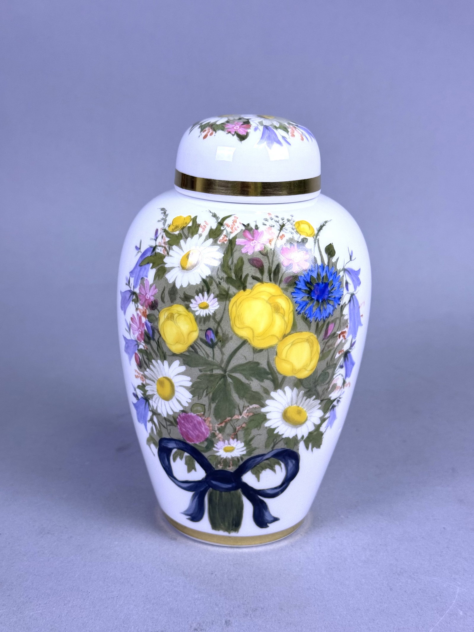 Nymphenburg Porcelain Lidded Floral Jar: This Nymphenburg porcelain jar features an ovoid form with a domed lid and is decorated with a hand-painted wildflower bouquet tied with a blue ribbon. The reverse and lid are further accented with sm