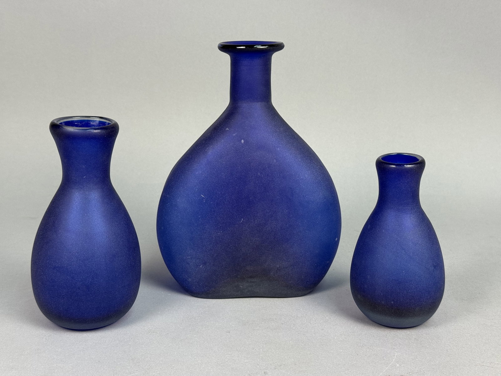 Trio of Cobalt Blue Satin Vases: A trio of hand-blown cobalt blue art glass vases featuring a uniform satin frosted finish. The collection consists of one large flattened bulbous vase with an elongated neck and two smaller pear-shape