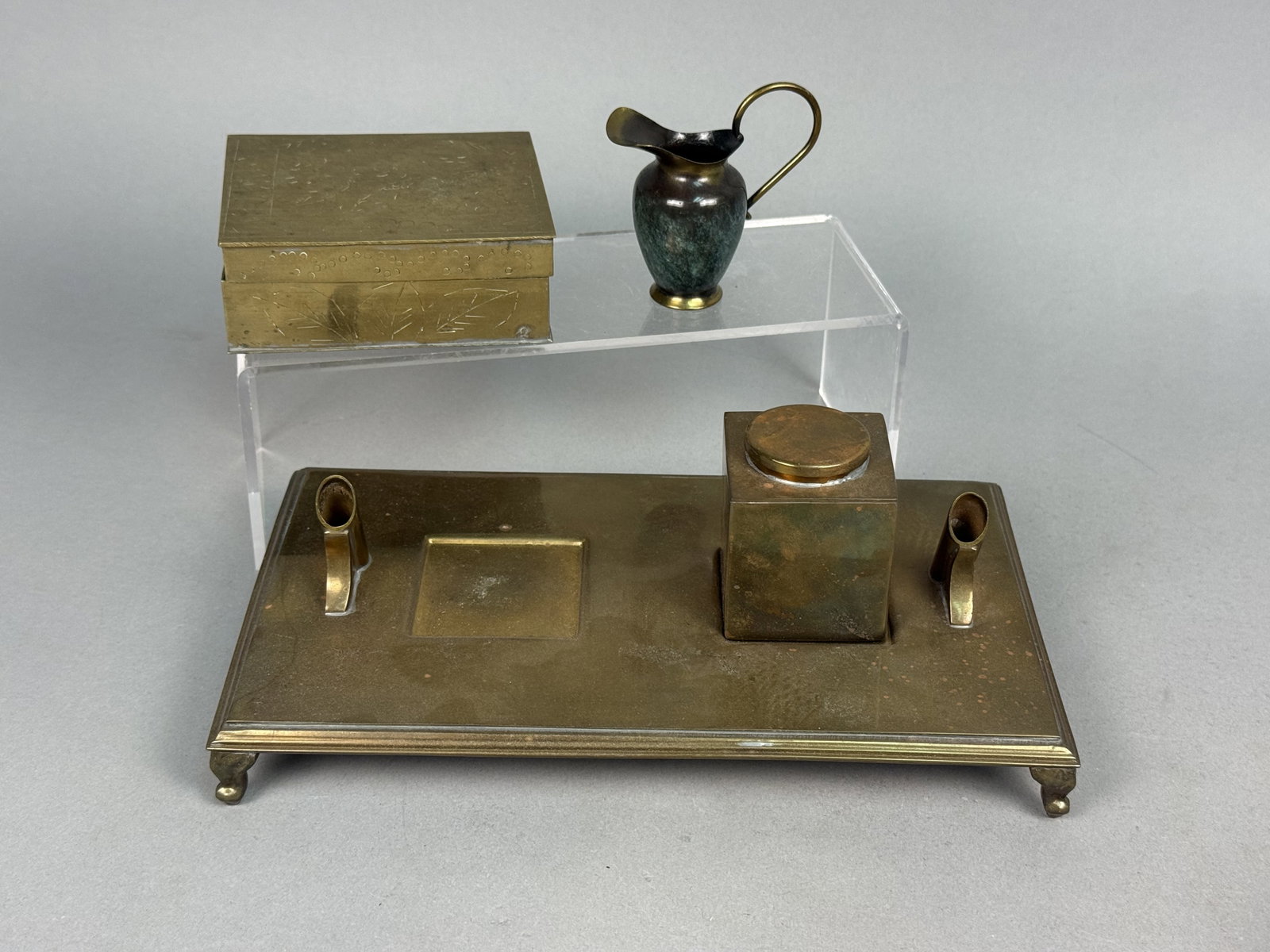 Collection of Brass Desk Accessories: This collection of brass desk accessories includes a Sarreid tray and inkwell, an etched box, and a miniature pitcher. The footed rectangular tray features dual angled pen holders and two recessed squ