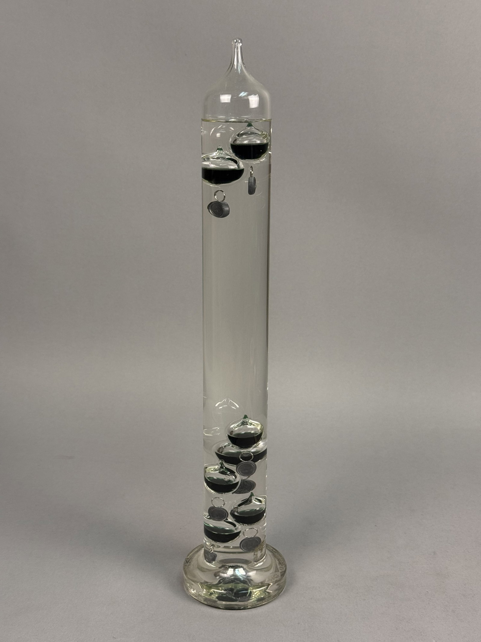 Large German Galileo Glass Thermometer: A large Galileo thermometer featuring a tall, sealed glass cylinder with a pulled-point tip and a flared circular pedestal base. The clear liquid-filled body contains eight glass buoyancy bulbs filled