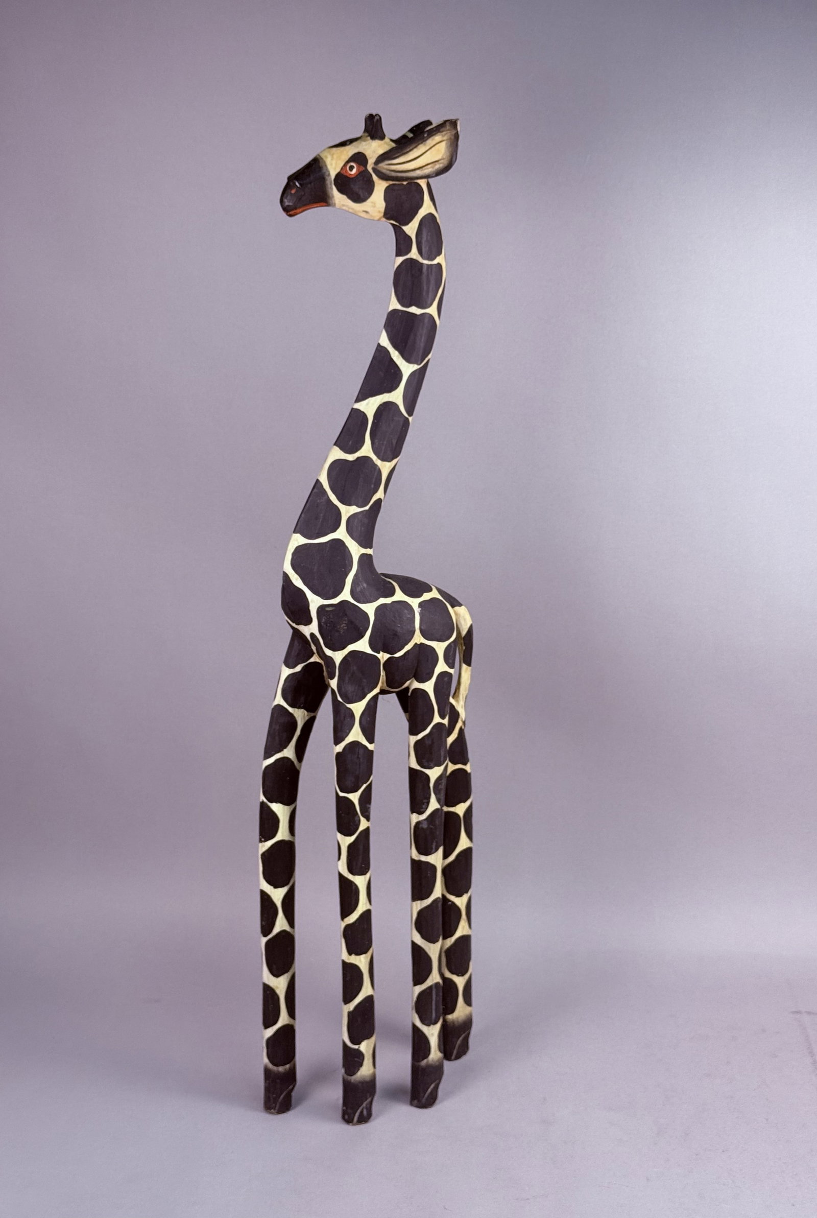 Hand-Carved Indonesian Painted Wooden Giraffe: This hand-carved wooden sculpture represents an elongated giraffe with a stylized upright posture. The surface is finished with a polychrome paint scheme featuring black irregular spots over a cream
