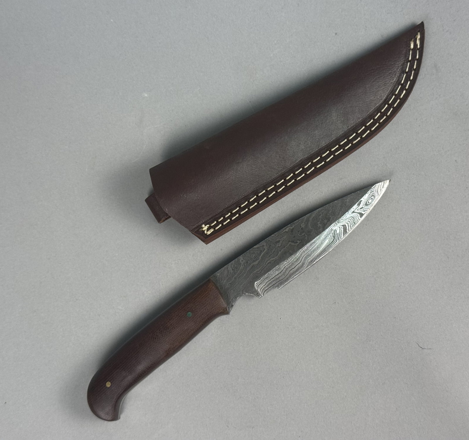 Damascus Steel Fixed Blade Knife: This fixed-blade hunting knife features a pattern-welded Damascus steel blade with a drop-point profile, thumb ramp, and finger choil. The full-tang construction is fitted with dark brown micarta hand